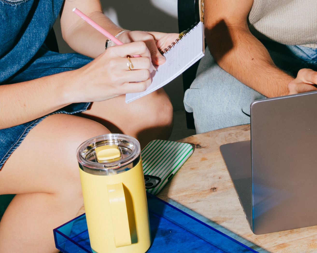 Two people sitting at a desk, one taking notes in a notebook, the other using a laptop. A yellow tumbler and mobile phone with green and white striped case are on the desk.