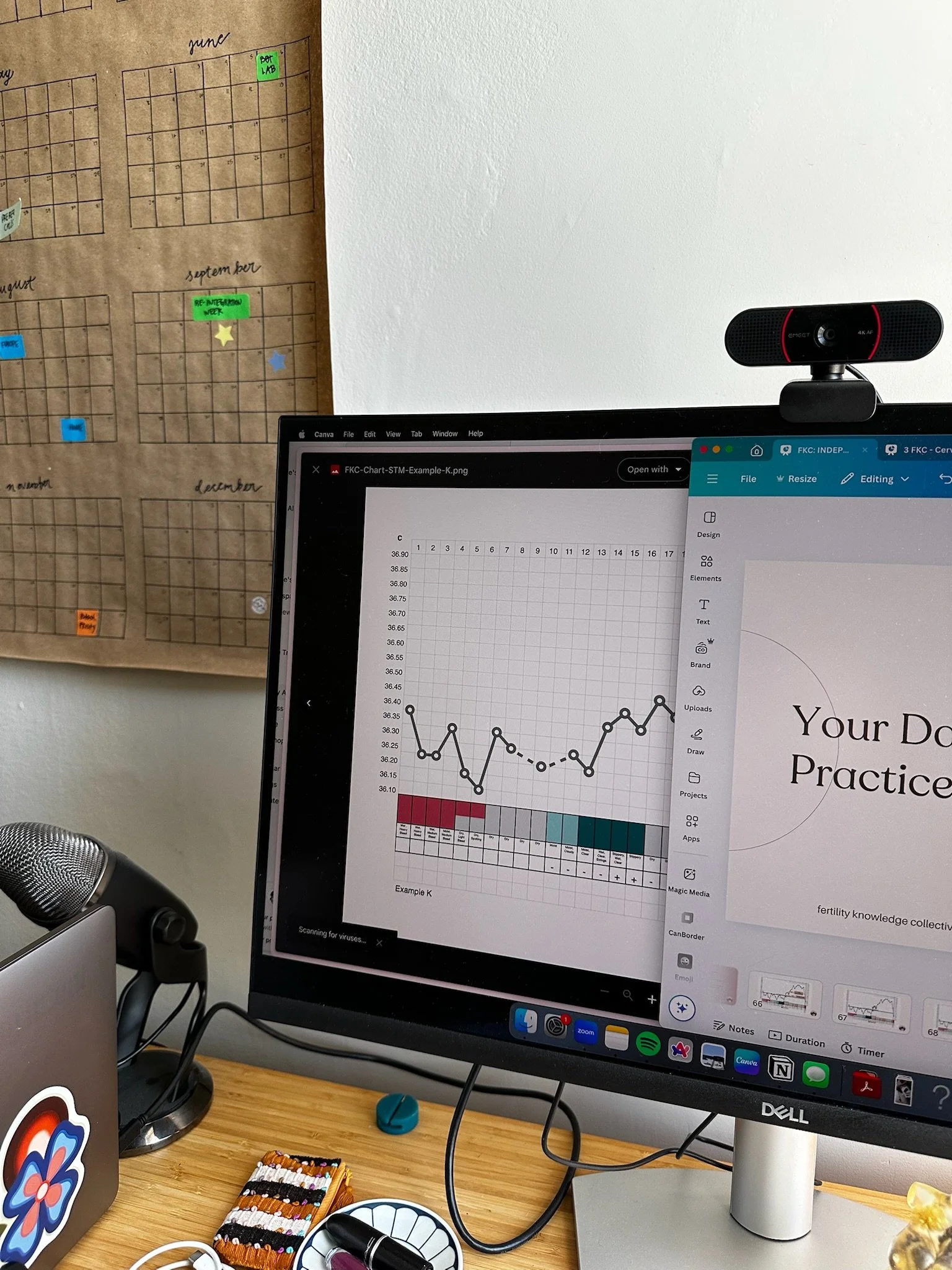Computer workstation with a monitor displaying a line graph and a file management window, a webcam on top of the monitor, a keyboard, and several stationery items on a wooden desk, with a bulletin board on the wall in the background.