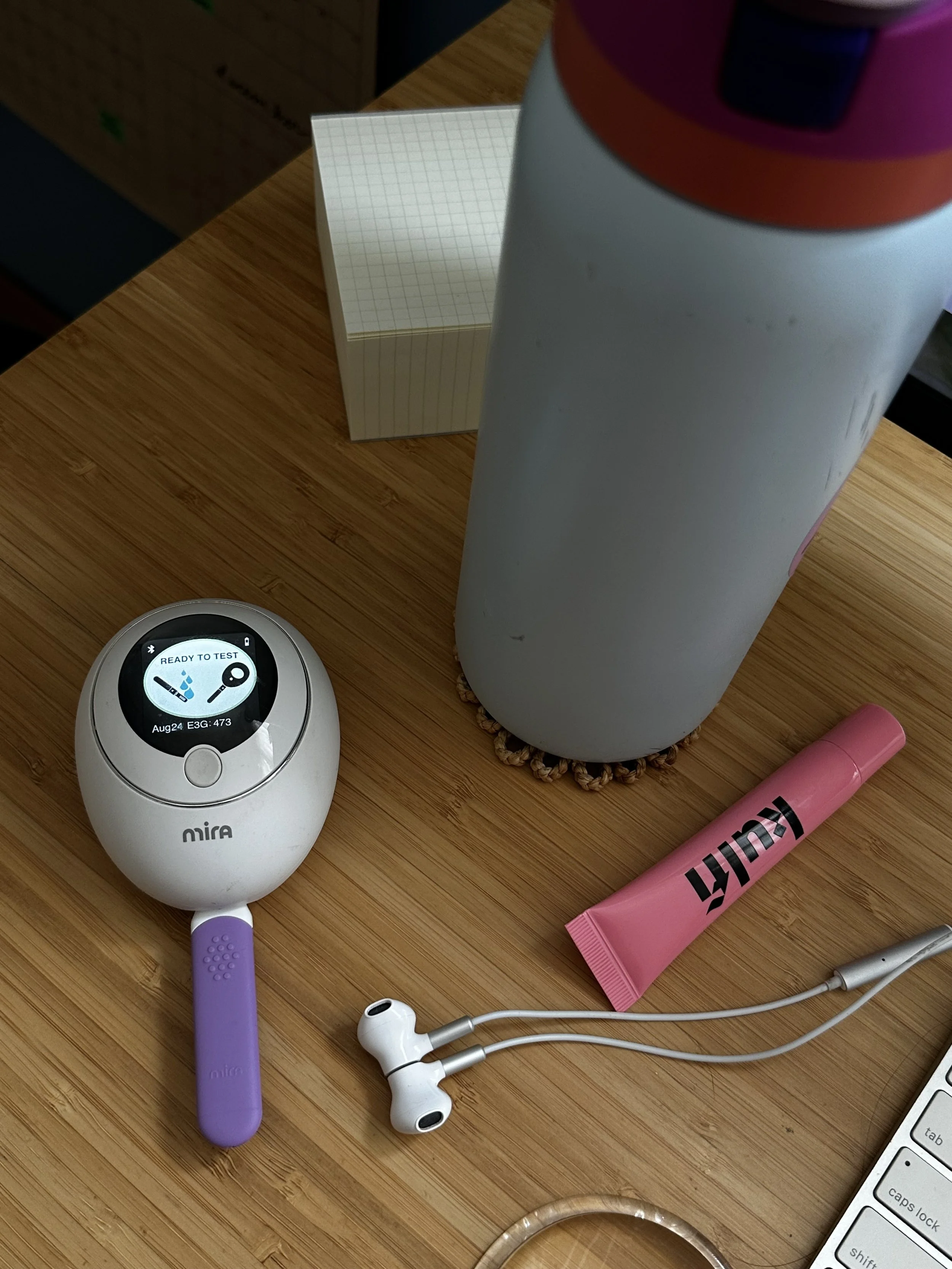 Various personal items on a wooden surface, including a purple toothbrush, white earbuds, a pink tube of lipstick, a white electronic device displaying temperature and humidity levels, and a cylindrical object with a grid paper note next to it.