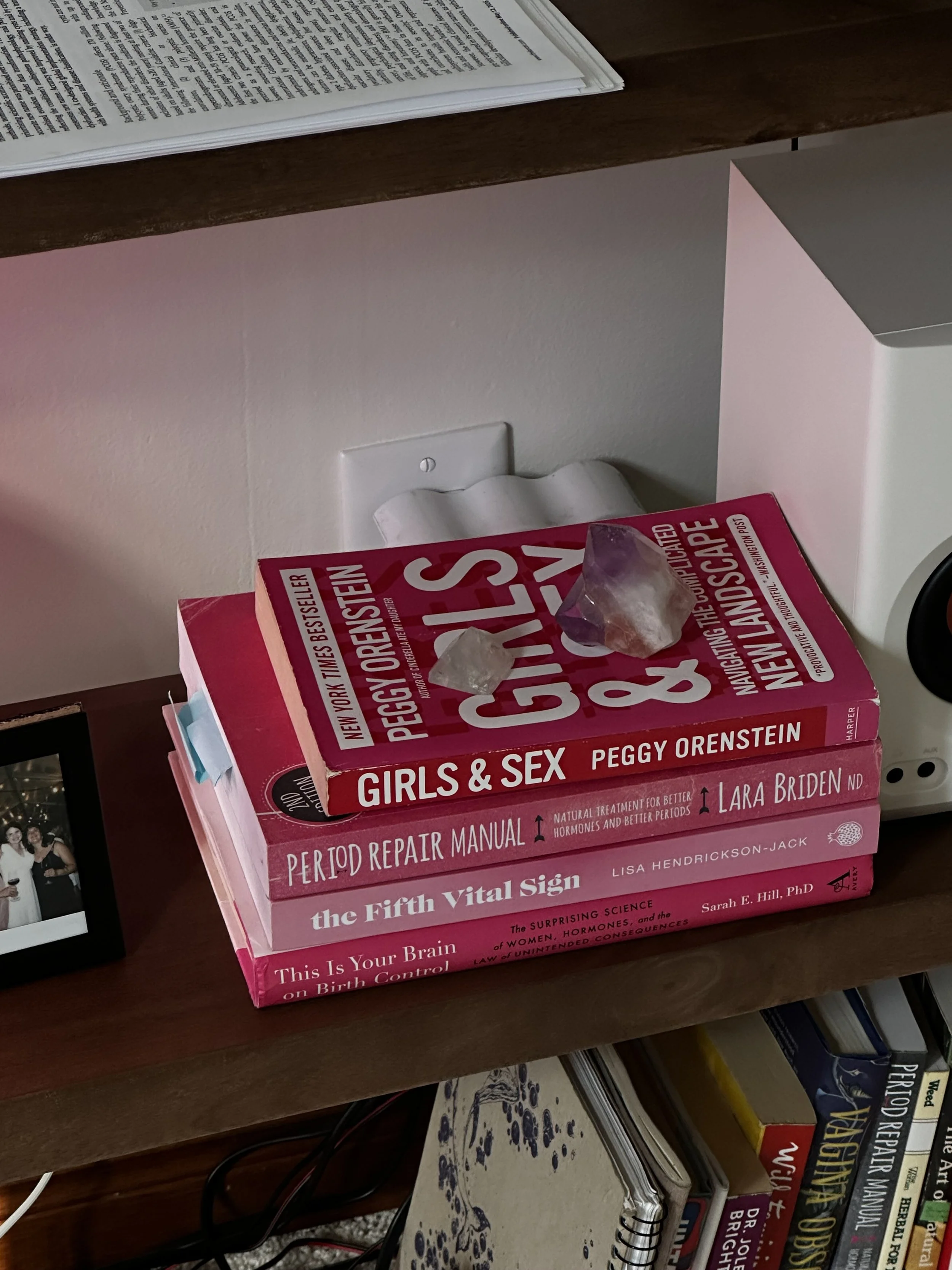 A stack of pink-colored books on a wooden surface, titled 'Girls & Sex' by Peggy Orenstein and other books about health, the brain, and reproductive health. An electrical outlet is visible behind the books. Part of a photo frame is seen at the bottom left corner.