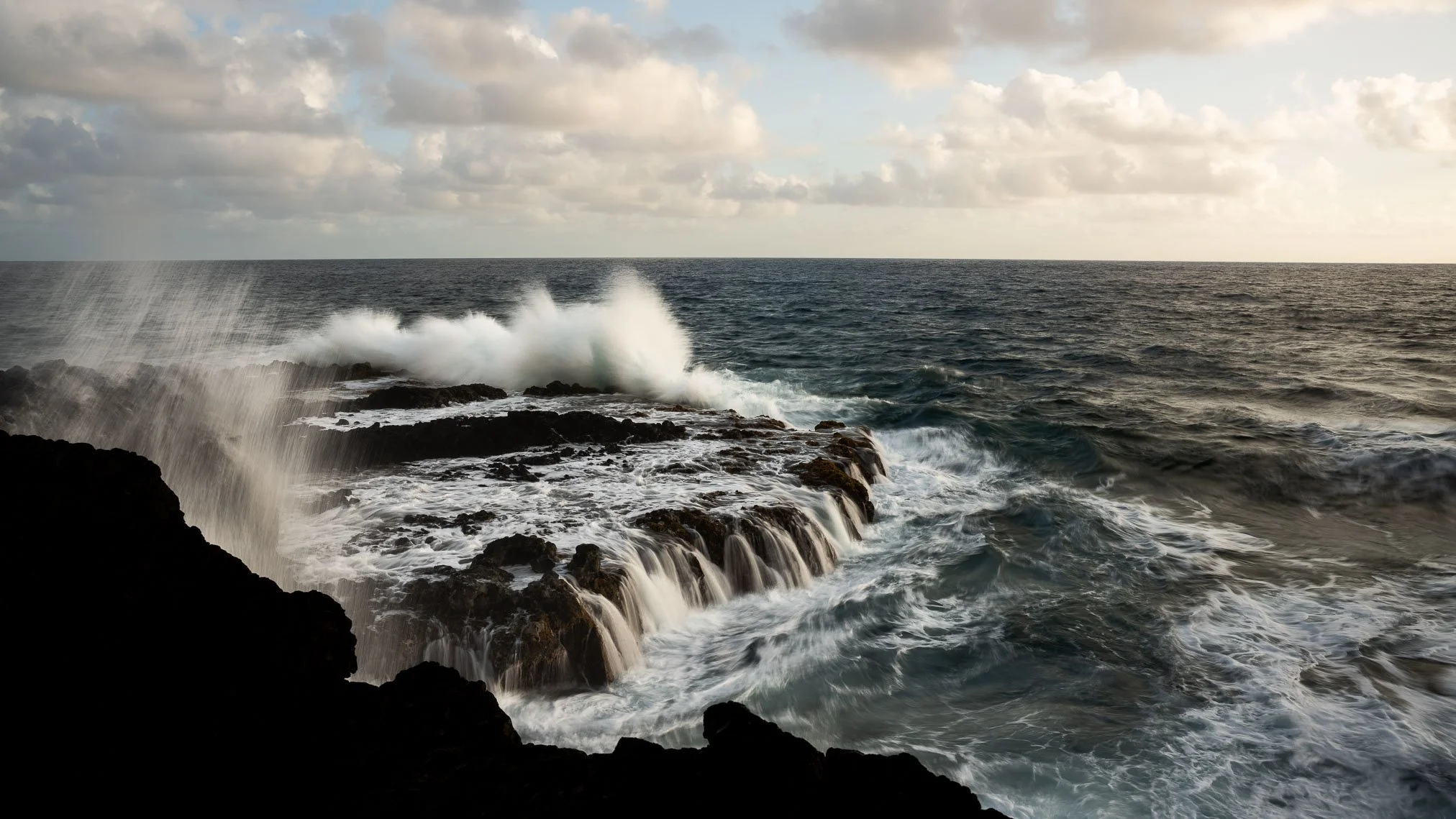 Ocean waves crashing against rocky shoreline in Kauai during cloudy sunset.