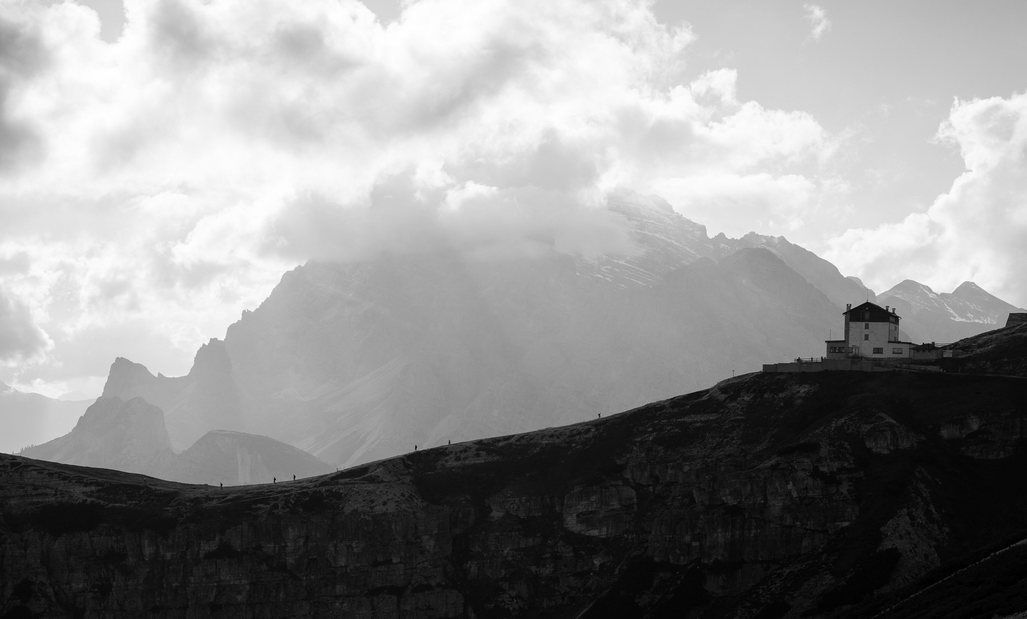 A black and white photo of a house perched on a hillside with mountains and cloudy sky in the background.