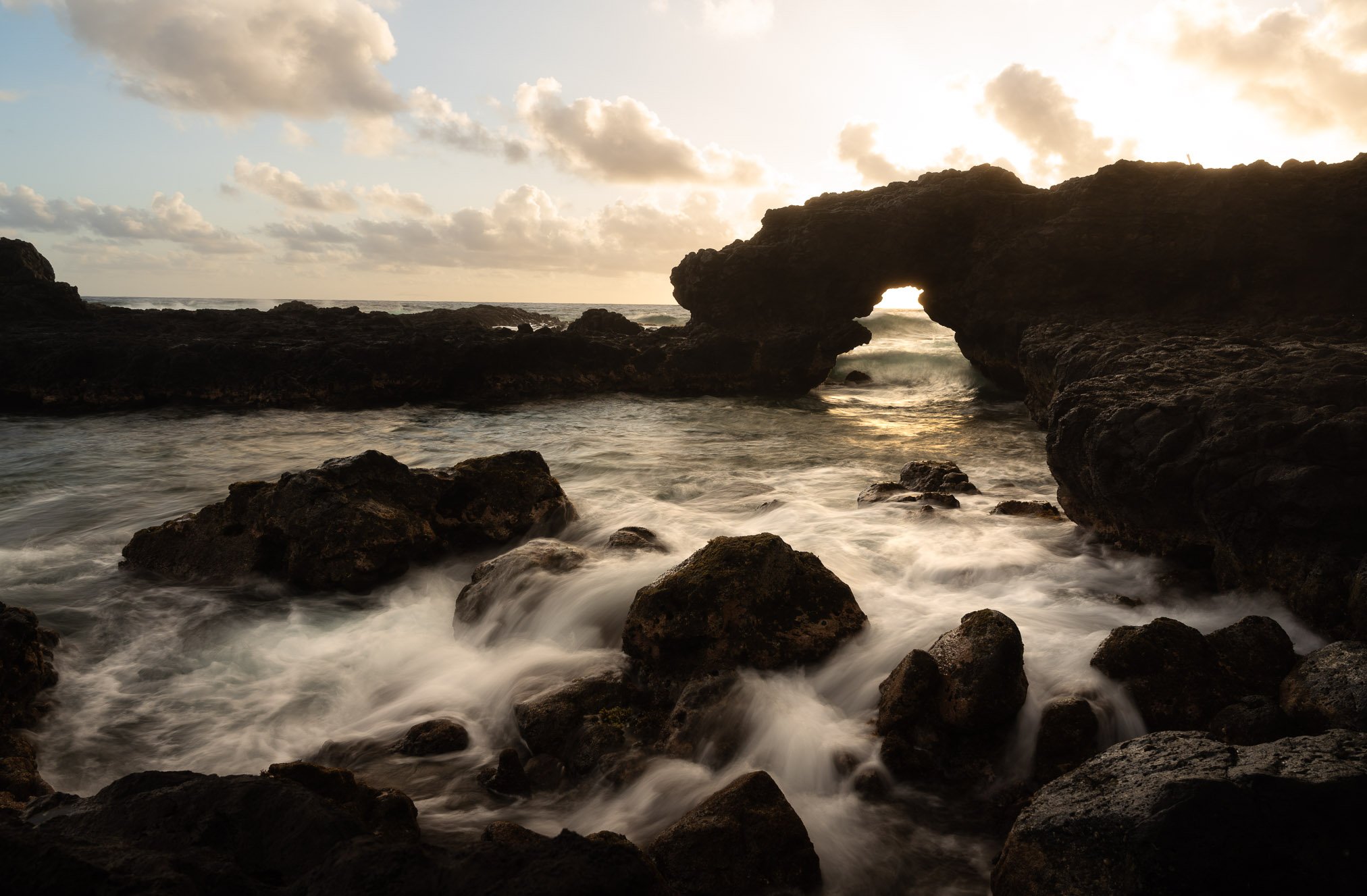 Sunset over rocky coastline in Kauai with a natural arch formation and ocean waves crashing against the rocks.