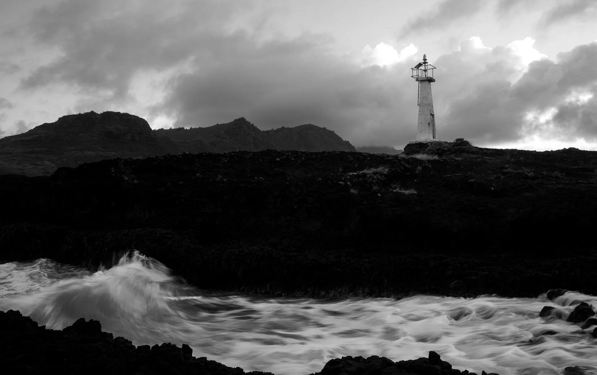Black and white photo of a rocky coastline in Hawaii with waves crashing on the shore, a lighthouse on a hill in the background, and clouds in the sky.