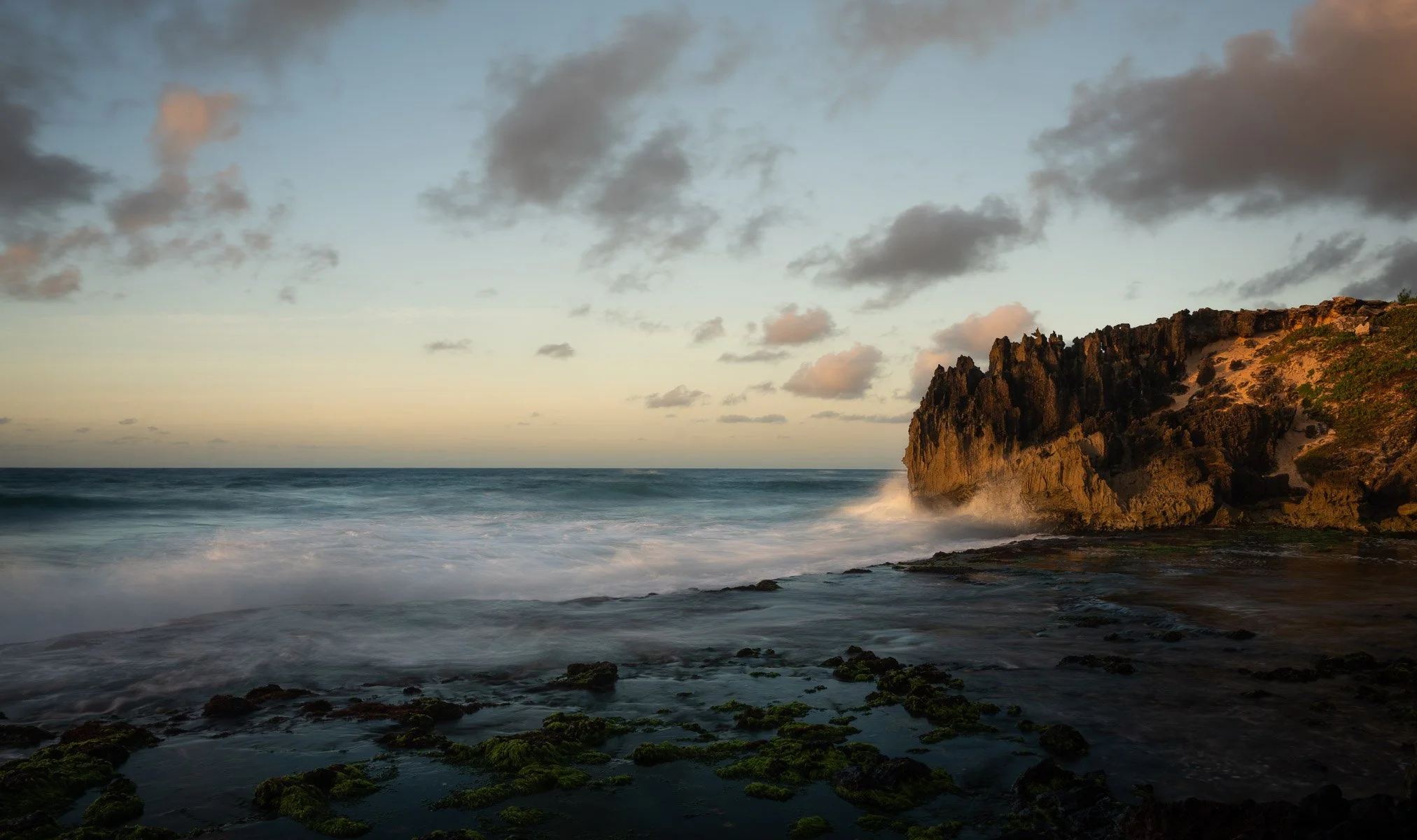 A scenic sunset over the ocean with waves crashing against a rocky cliff on the right, and some moss-covered rocks in the foreground.