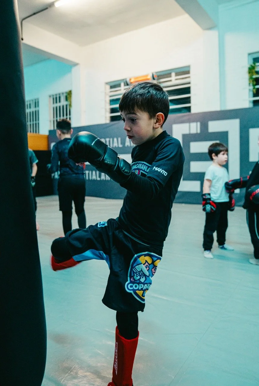 A young boy practicing martial arts on a punch bag in a gym, wearing boxing gloves, shin guards, and sports attire, with other children in the background.