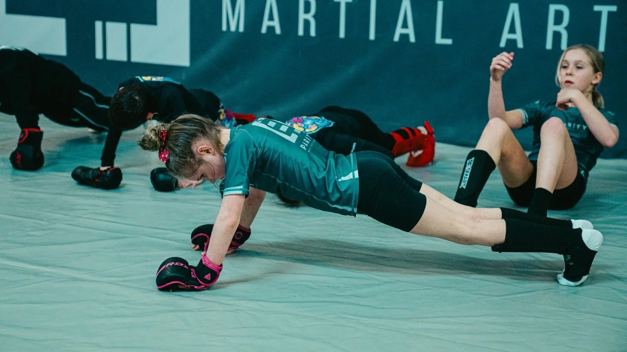 Young girls practicing martial arts strength and conditioning on a padded floor in a training studio with a large banner in the background.