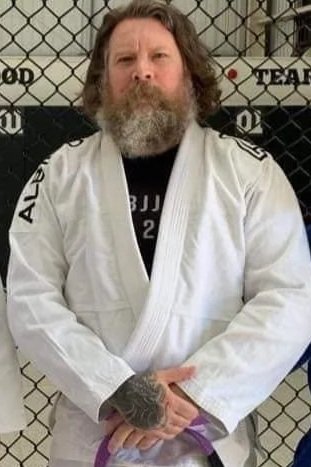 A man with long hair and a beard wearing a white jiu-jitsu gi standing in front of a chain-link fence.