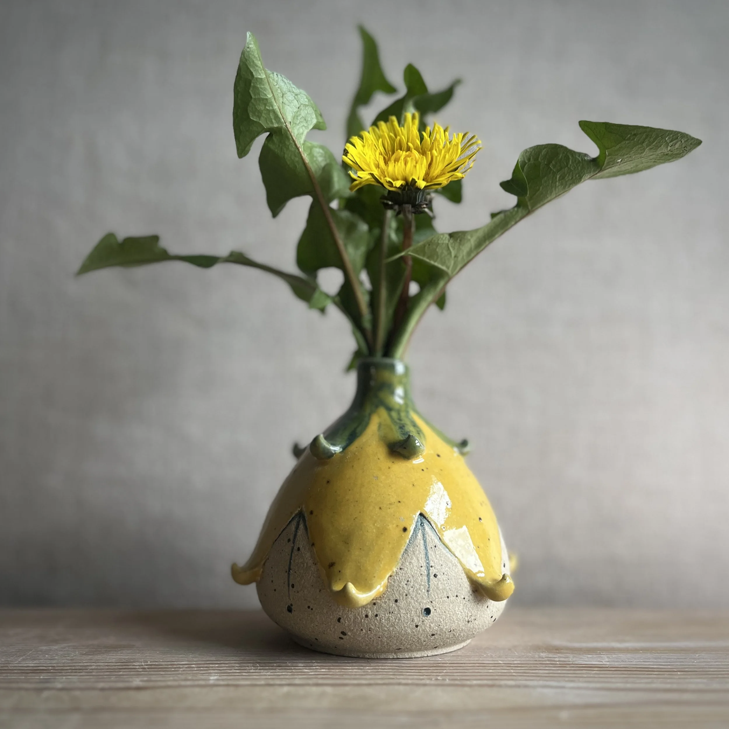 Yellow and green vase in the shape of an upside-down flower head with a dandelion in it on a wooden surface