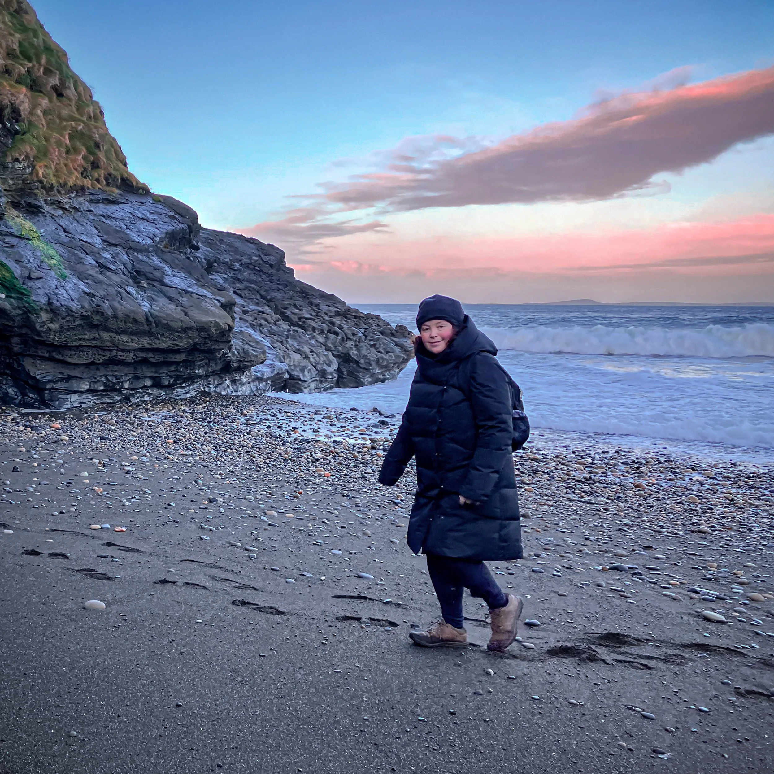 Woman in a black coat walking on a rocky beach with cliffs and ocean waves.