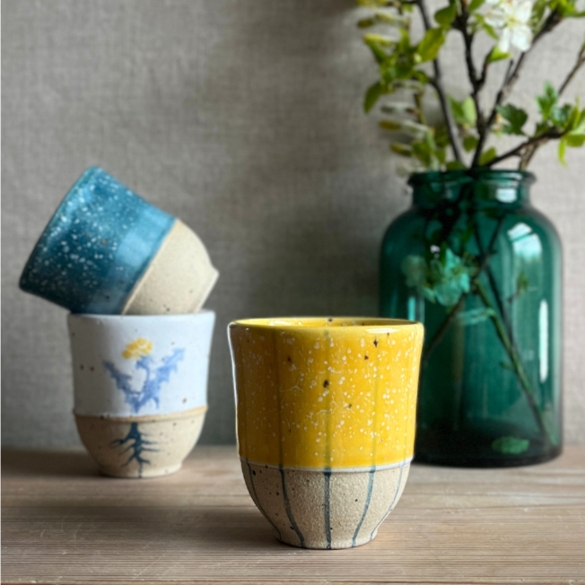 Three small ceramic cups, one yellow, one blue and one with a dandelion print on a wooden surface with a green vase and leafy plant in the blurred background.