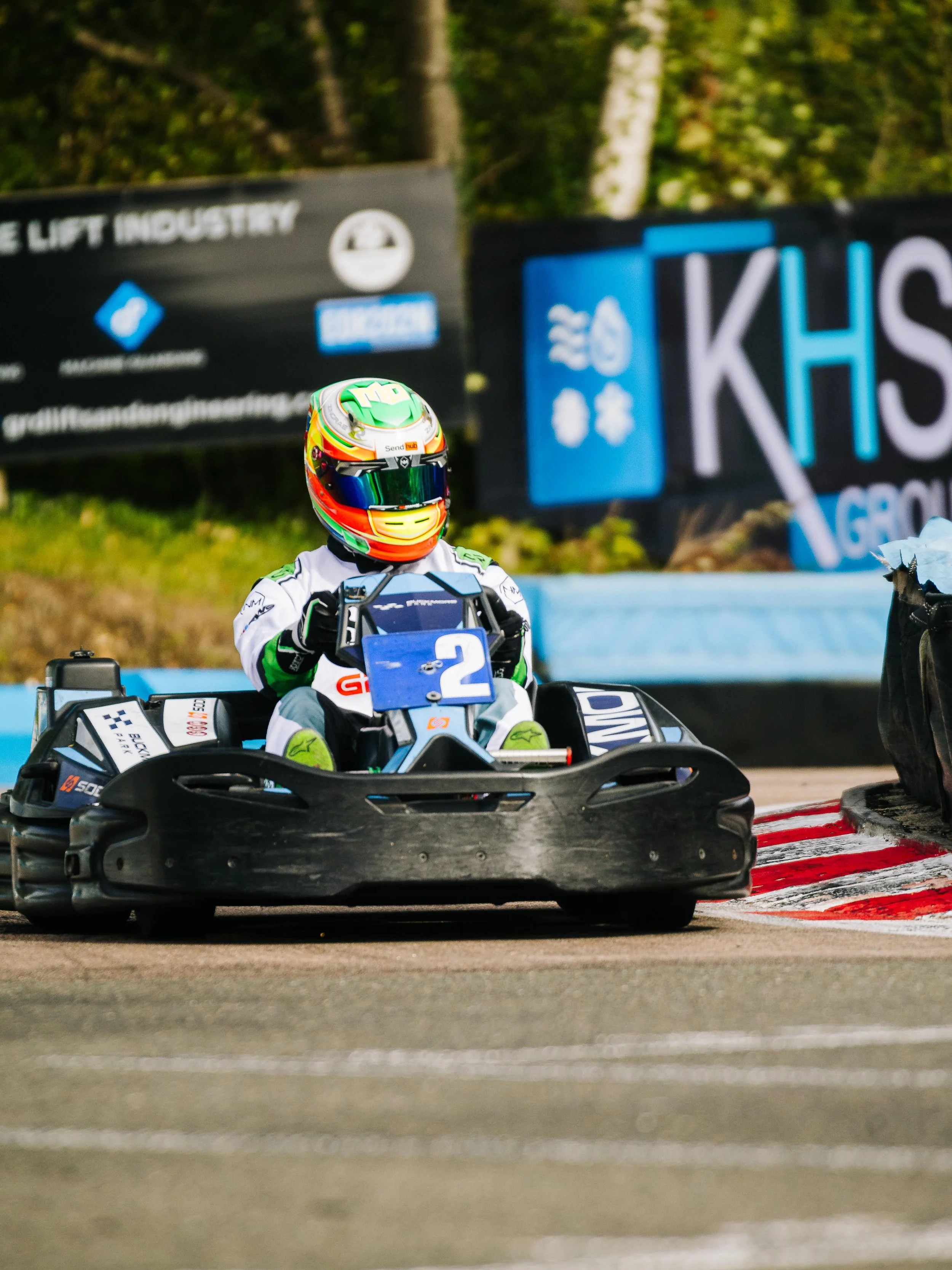 A young person racing go-kart on a track, wearing a colorful helmet and white racing suit, with a blue number 2 on the front of the go-kart.
