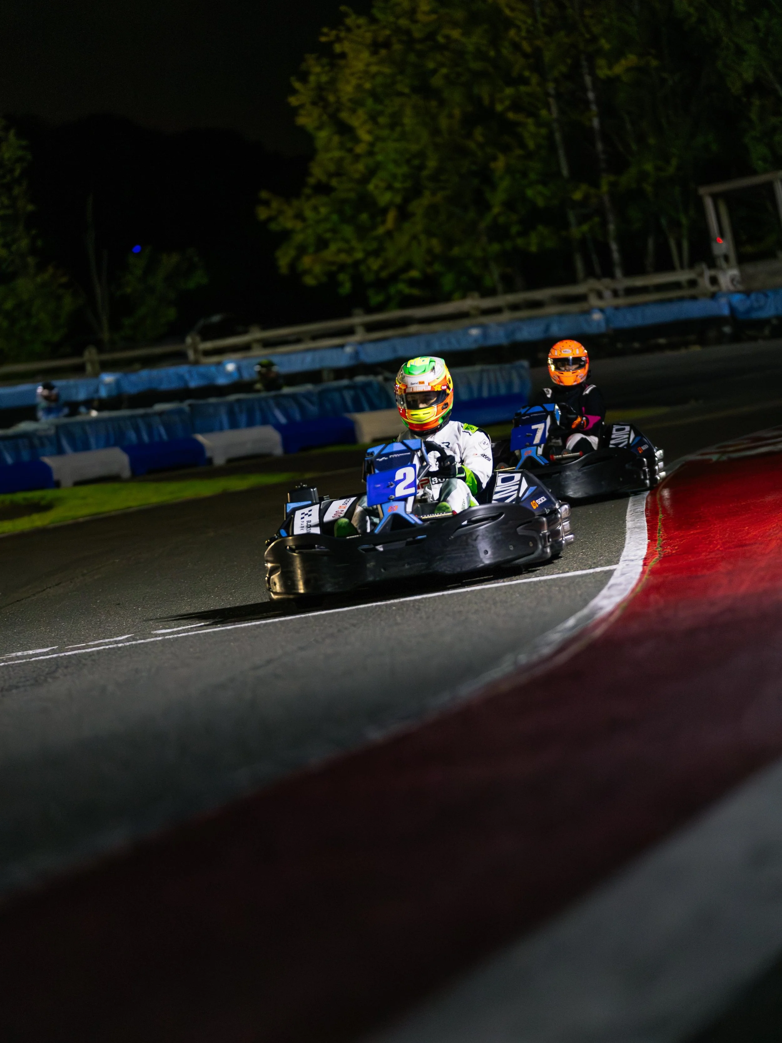 Nighttime go-kart race with two drivers wearing helmets and racing suits, navigating a curve on a racetrack.