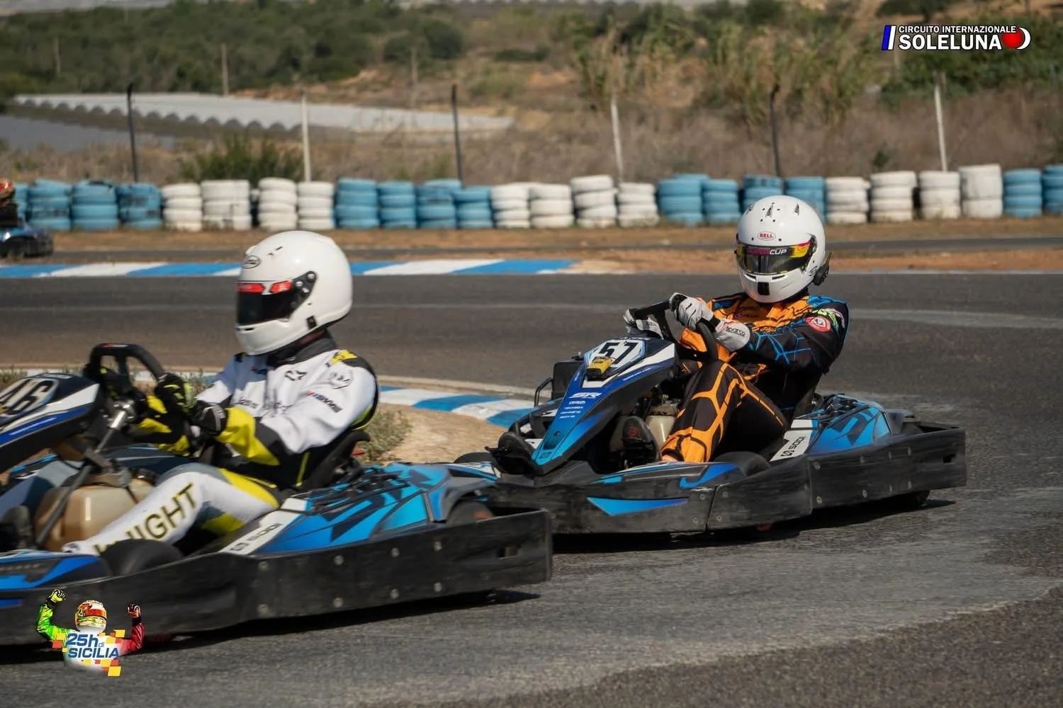 Two go-kart racers in helmets and racing suits on a race track, with a background of tire barriers and a hilly landscape, participating in an event at Circuito Internazionale Solleuna.
