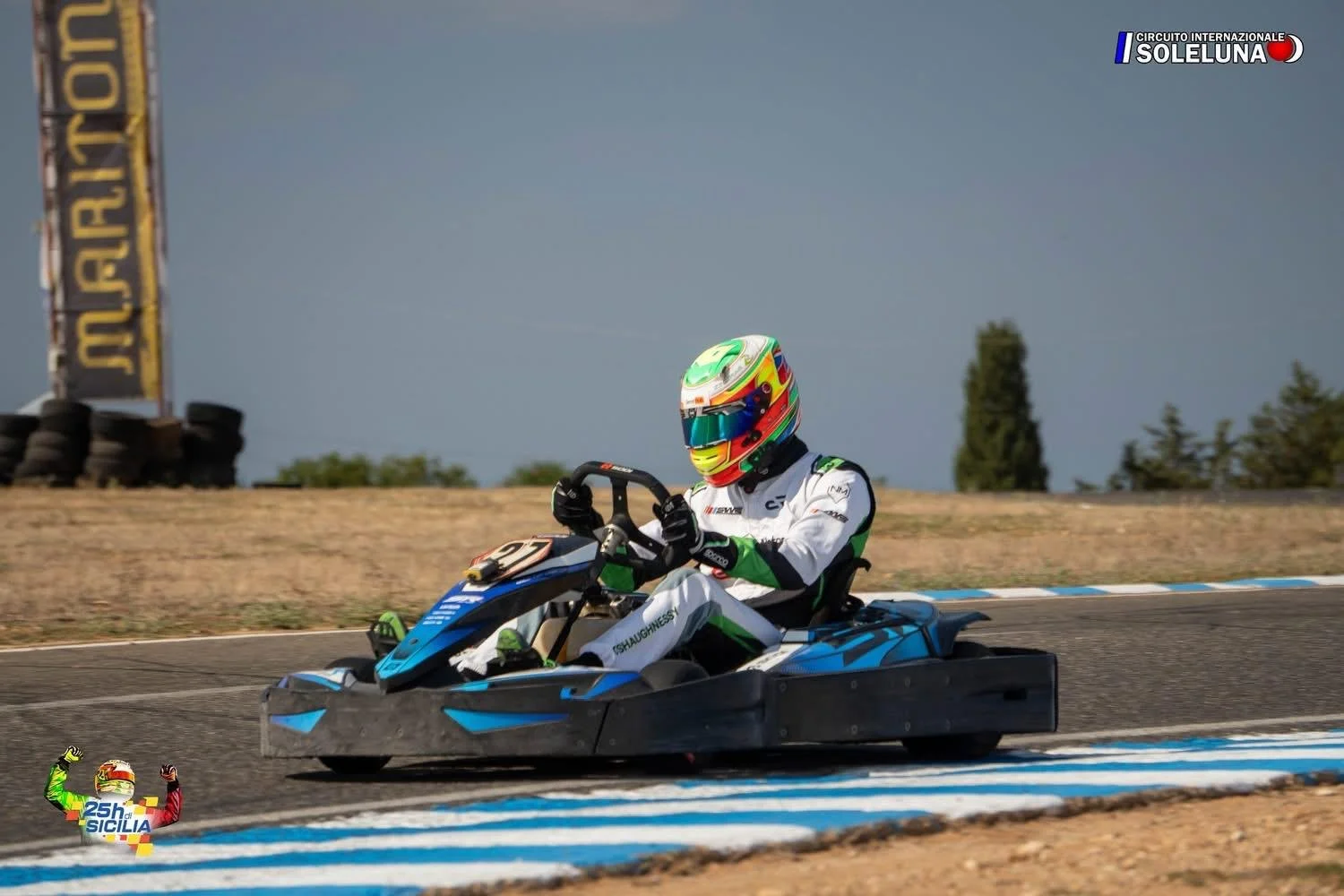 Race car driver in full gear driving a go-kart on a race track, wearing a colorful helmet and white racing suit with black and green accents, during the 25th Sicily Karting event at Circuito Internazionale SolLuna.