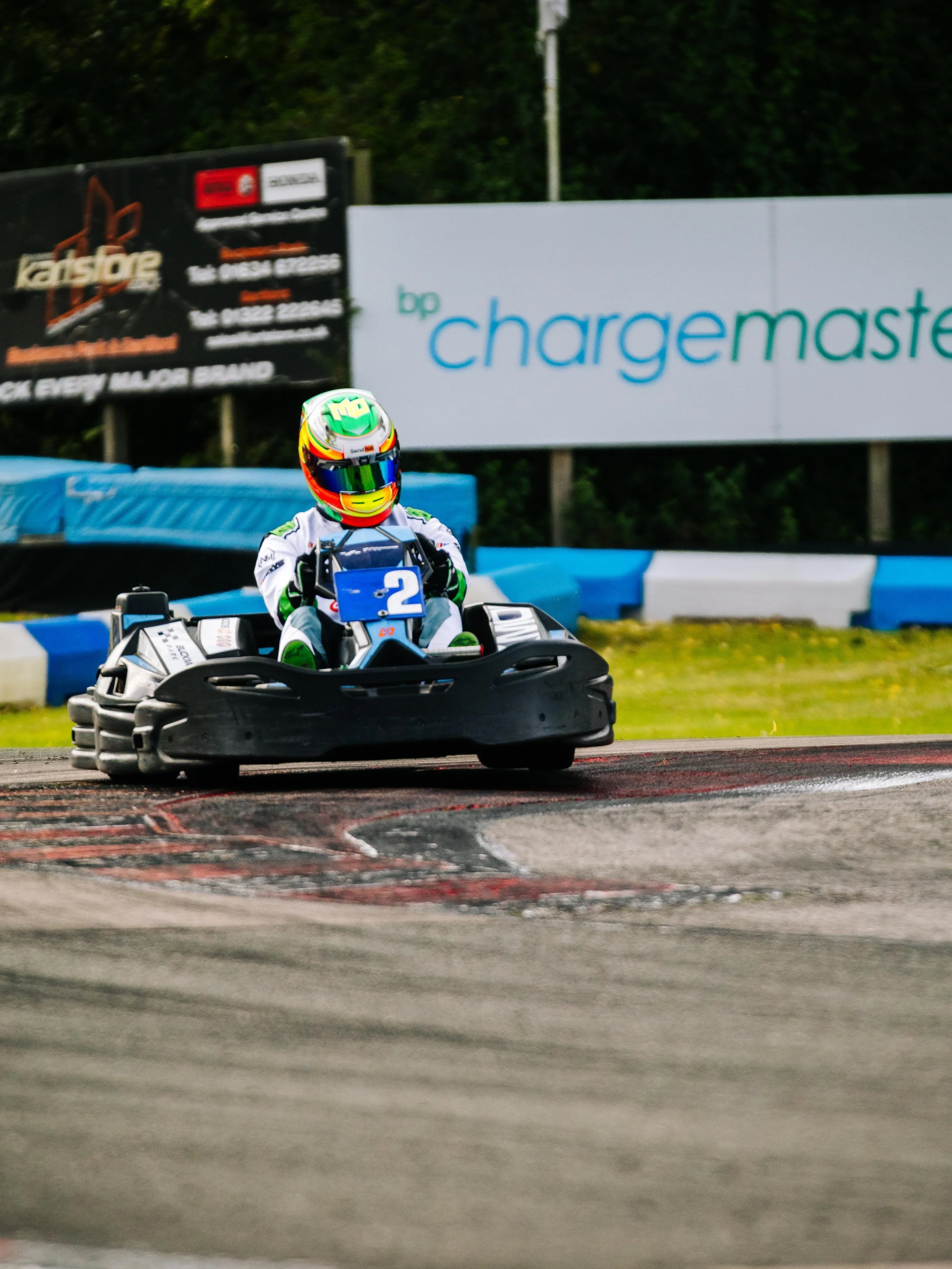 Go-kart racer wearing a colorful helmet and white racing suit, steering a go-kart on a race track with advertisements in the background.