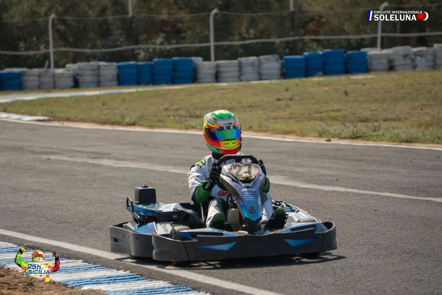 A go-kart racer wearing a colorful helmet and racing suit on a race track, with blue and white tire barriers in the background, during the SolLUNA International Circuit event.