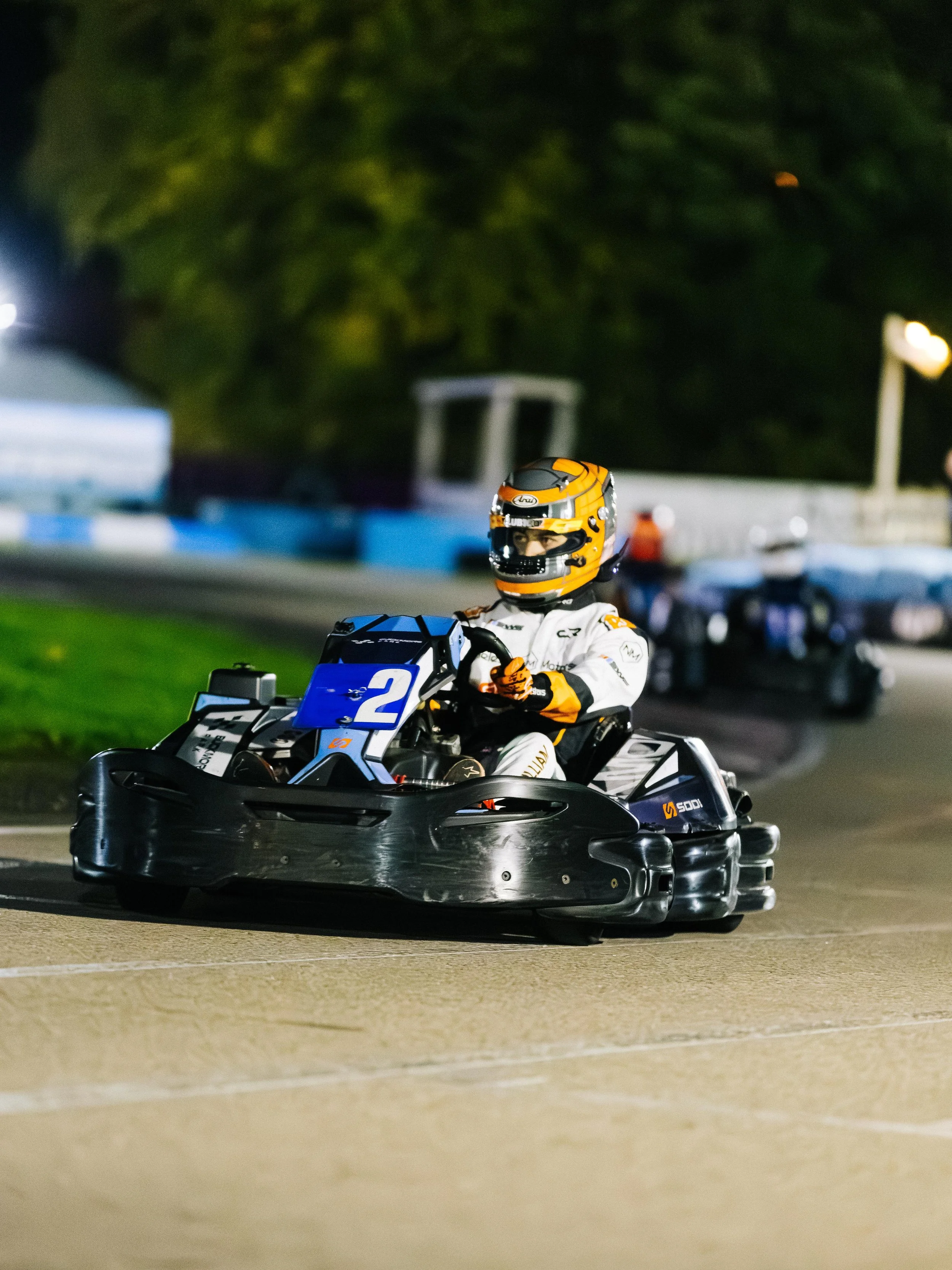 A person in a white racing suit and yellow helmet driving a go-kart on a nighttime race track.