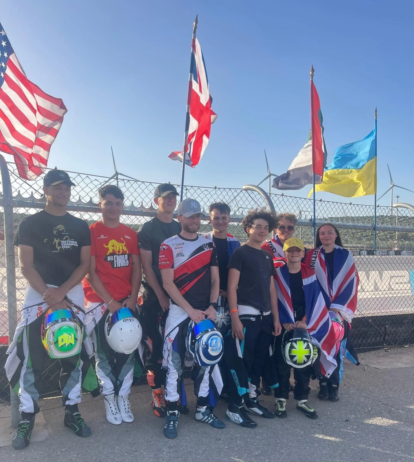 Group of young race car drivers and their team standing in front of a fence with national flags, holding helmets and wearing race gear, under a clear blue sky.