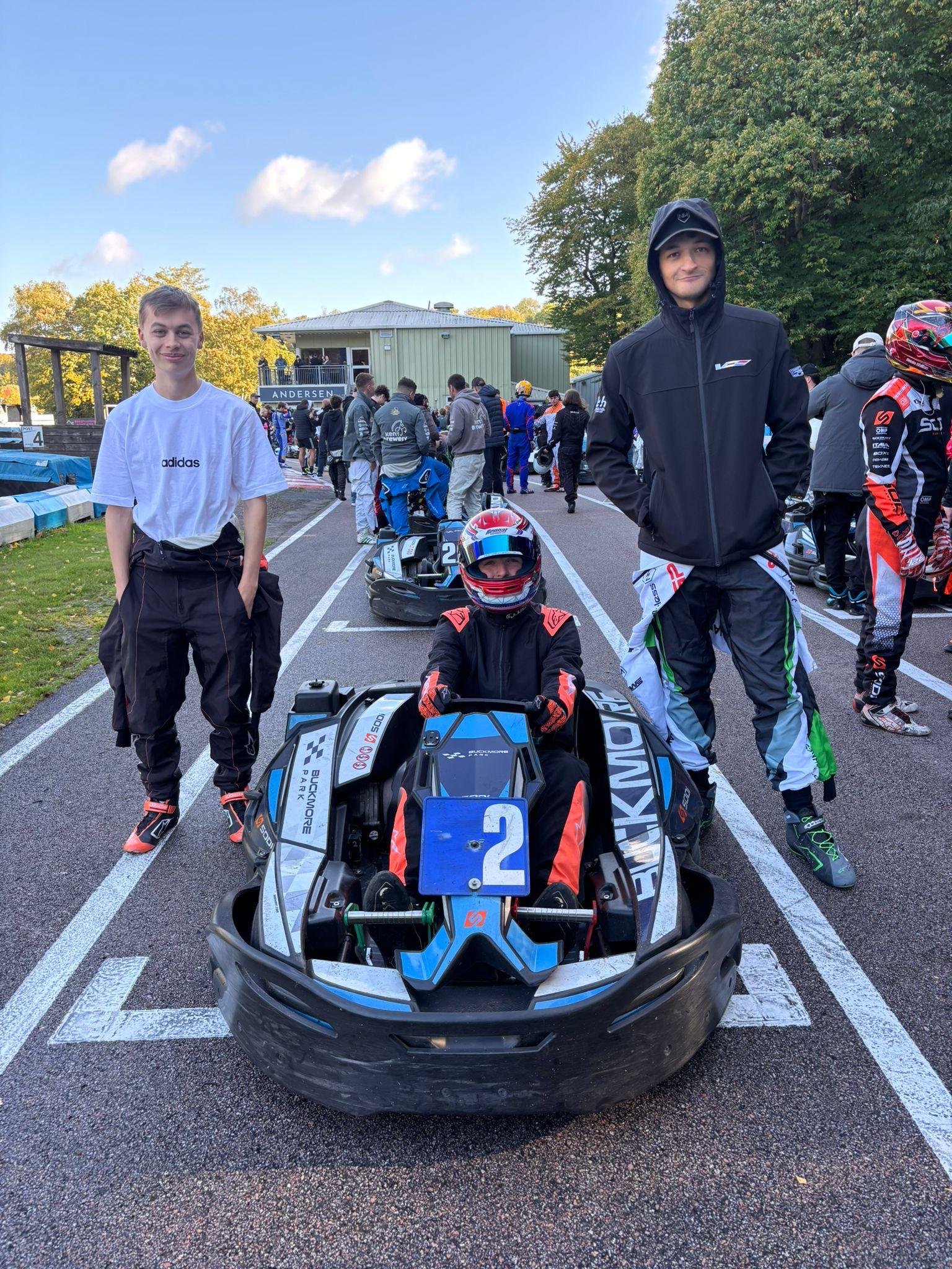 Young race car driver in a racing suit and helmet sitting in a go-kart labeled with the number 2, flanked by two taller individuals, in a race track paddock area with people and race cars in the background.