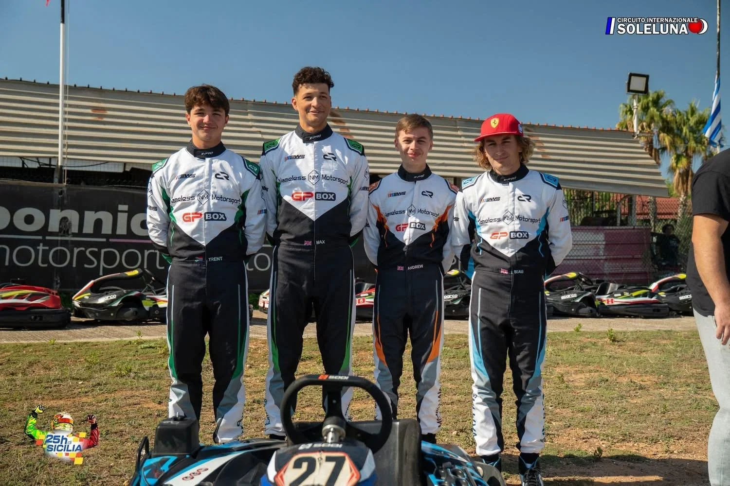 Four young race car drivers in racing suits standing on grass with go-karts behind them at a racetrack, posing for a photo during daytime at Circuito Internazionale Sol Luna, Sicily.