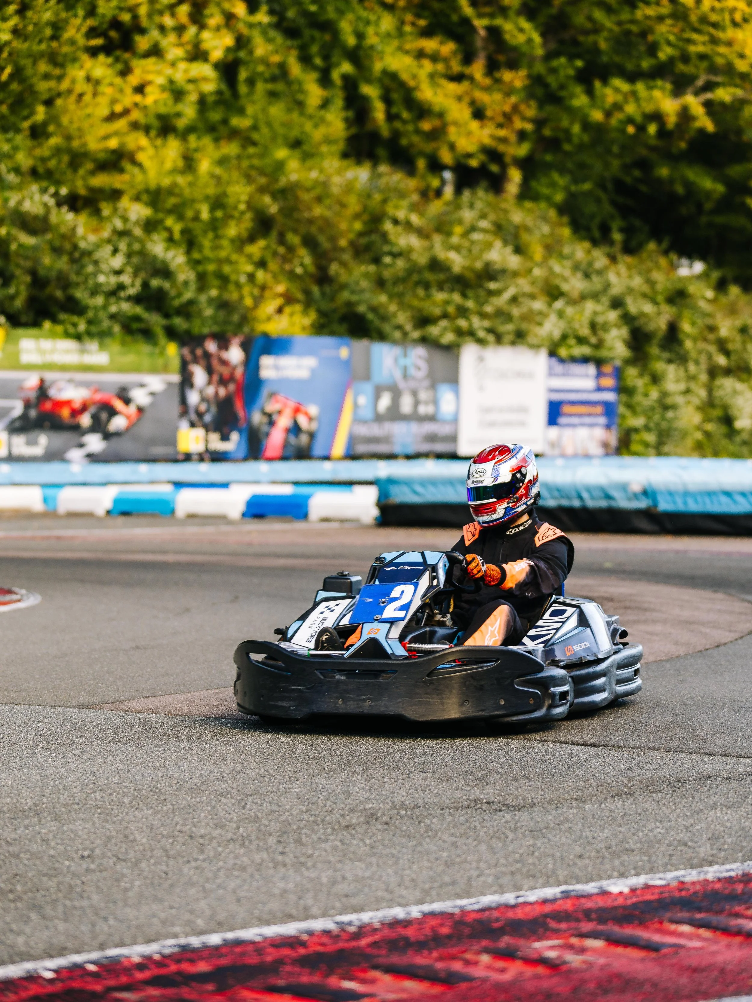 Person in a racing go-kart on a race track, wearing a helmet and racing gear, with a background of trees and advertisements.