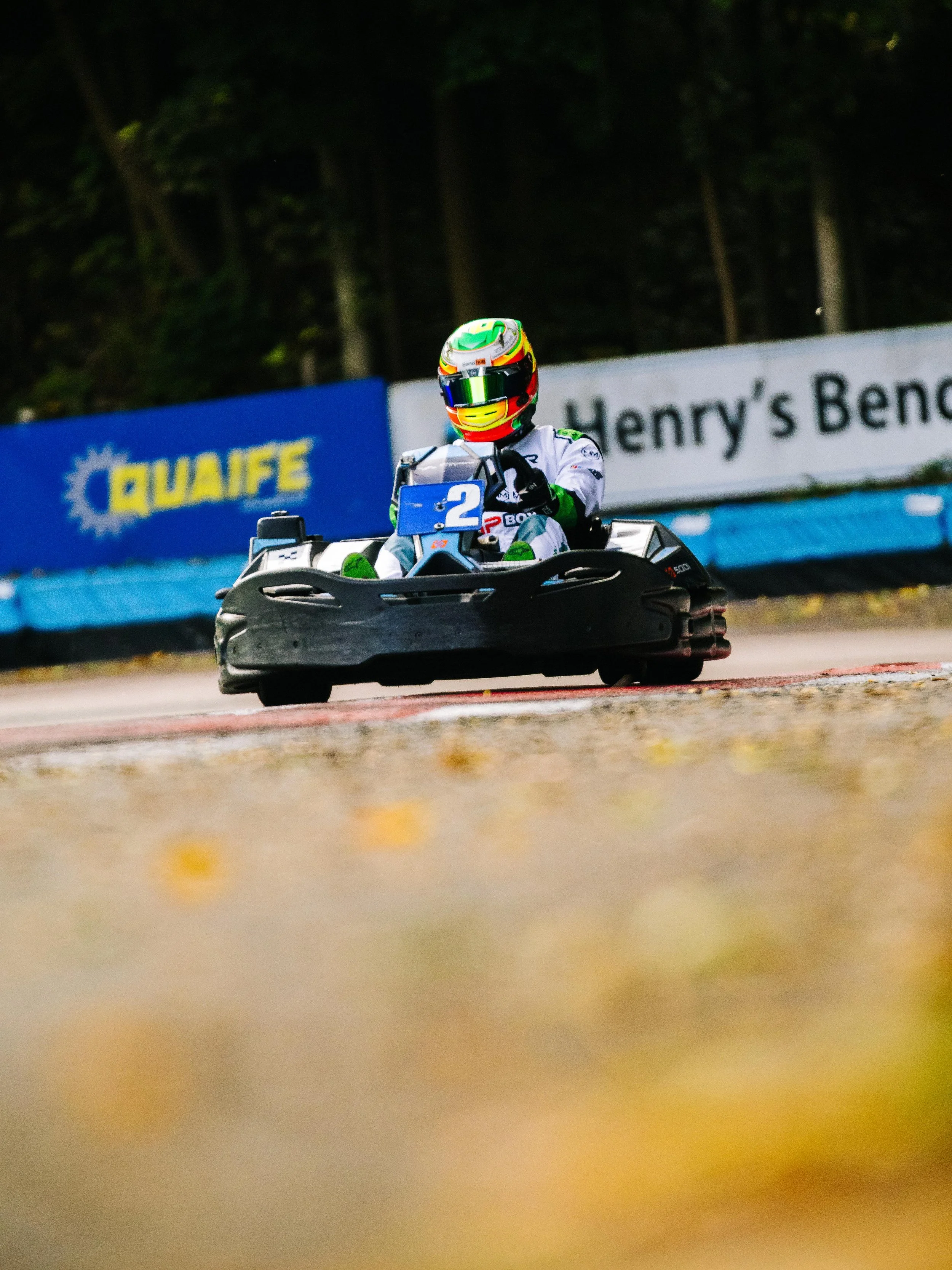 A person racing a go-kart on a track, wearing a white racing suit and a multicolored helmet, with banners and trees in the background.