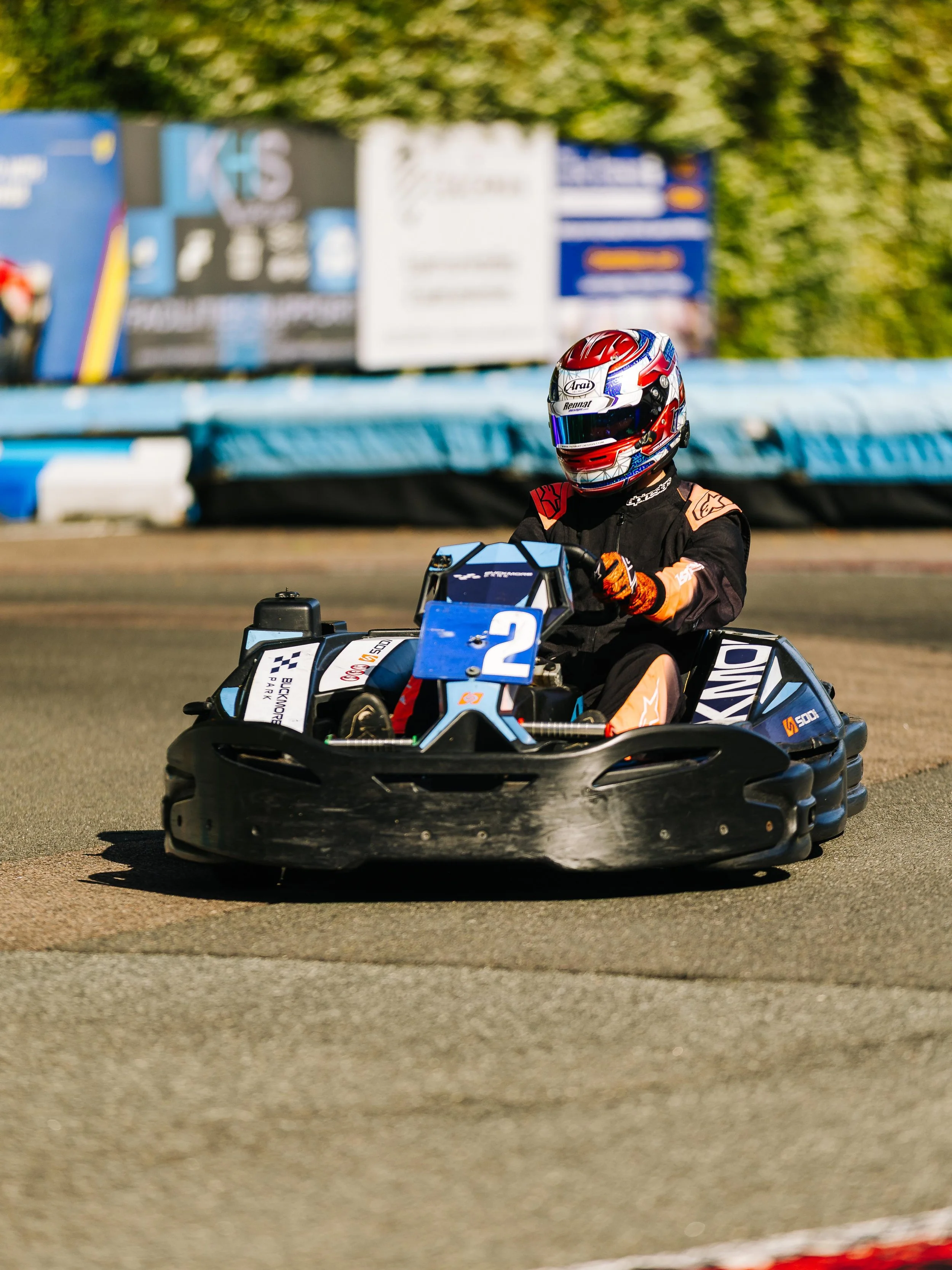 A person in racing gear driving a blue go-kart with the number 2 on it on a race track.