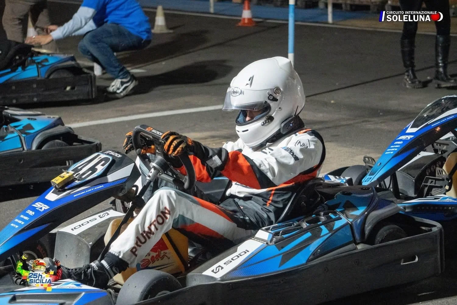 A race car driver in a white helmet and racing suit sitting in a blue and black go-kart at a racetrack, with other people and go-karts in the background.