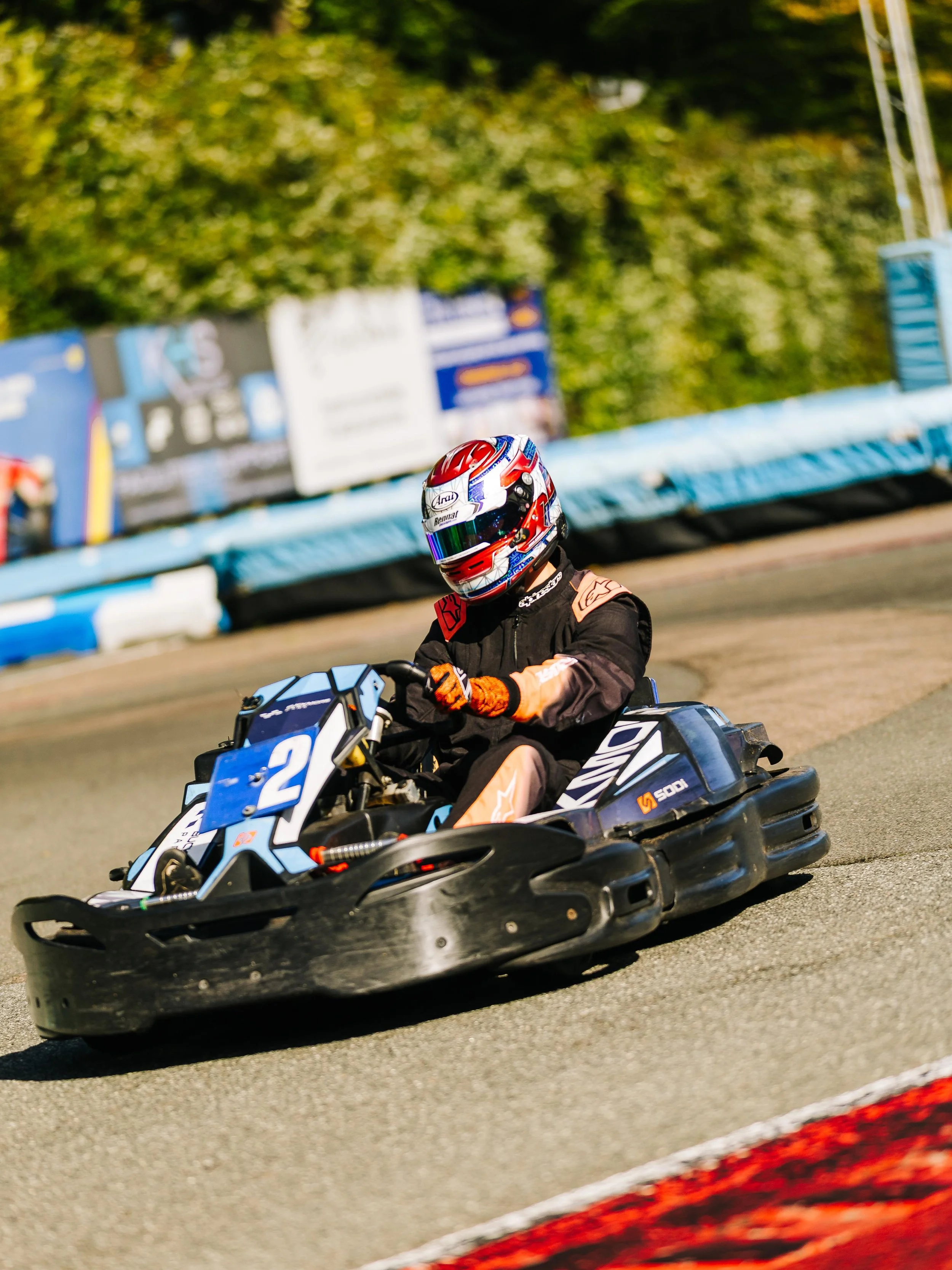 A person in racing gear, including a helmet, driving a go-kart on a race track during the daytime.