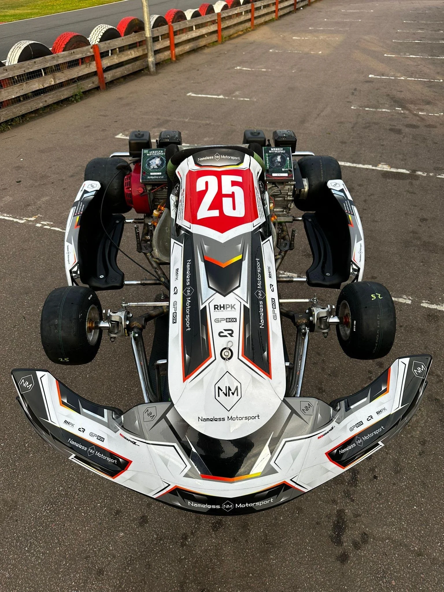 Race car with number 25, black, white, red, and gray decal design, parked on asphalt, with tire barrier in background.