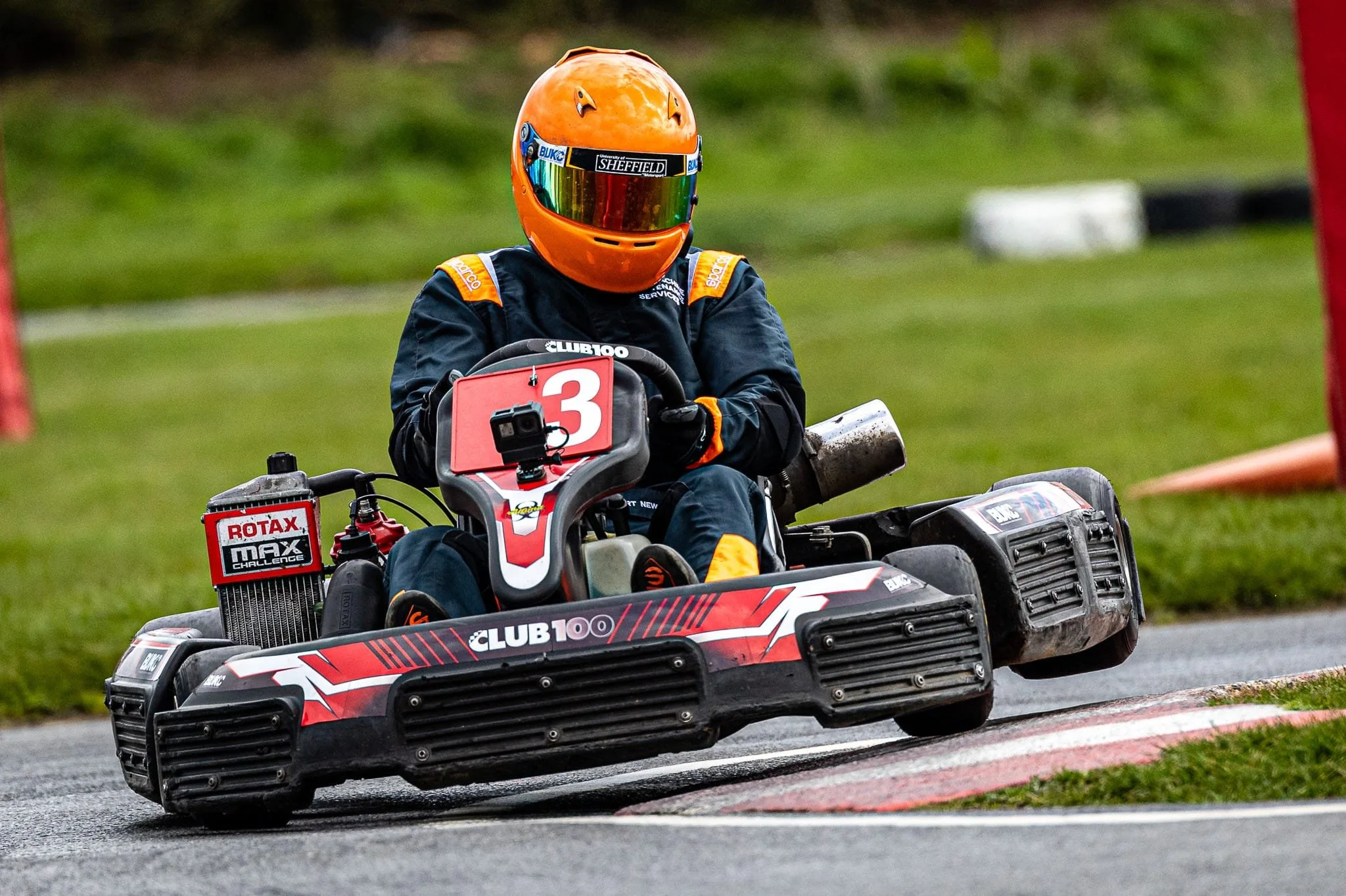 A go-kart driver wearing an orange helmet and black racing suit, navigating a corner on a race track. The go-kart is black and red with the number 3 on the front and various sponsor stickers.