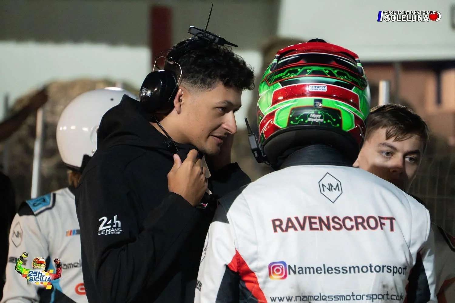 Race car drivers and team members preparing in the pit area during a race at Circuito Internazionale Solleuna, with one person adjusting equipment on another driver wearing a helmet and team jacket.