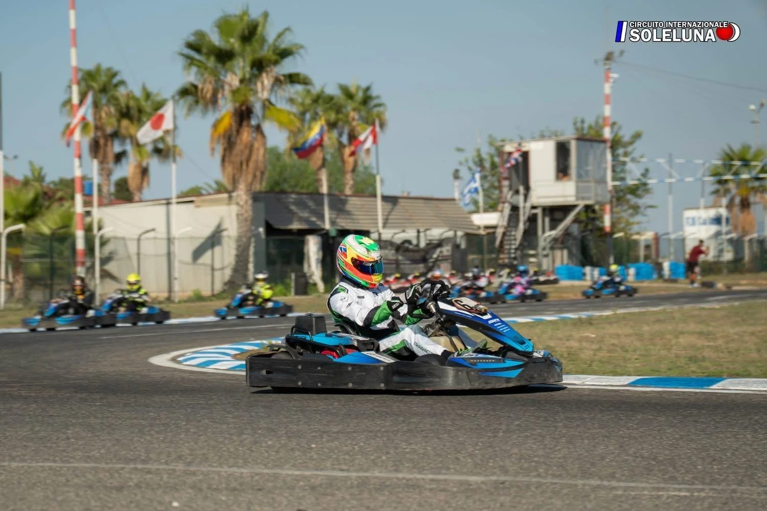 Go-kart race with a driver wearing a white racing suit and colorful helmet, in a sharp turn on a race track, with palm trees and flags in the background.