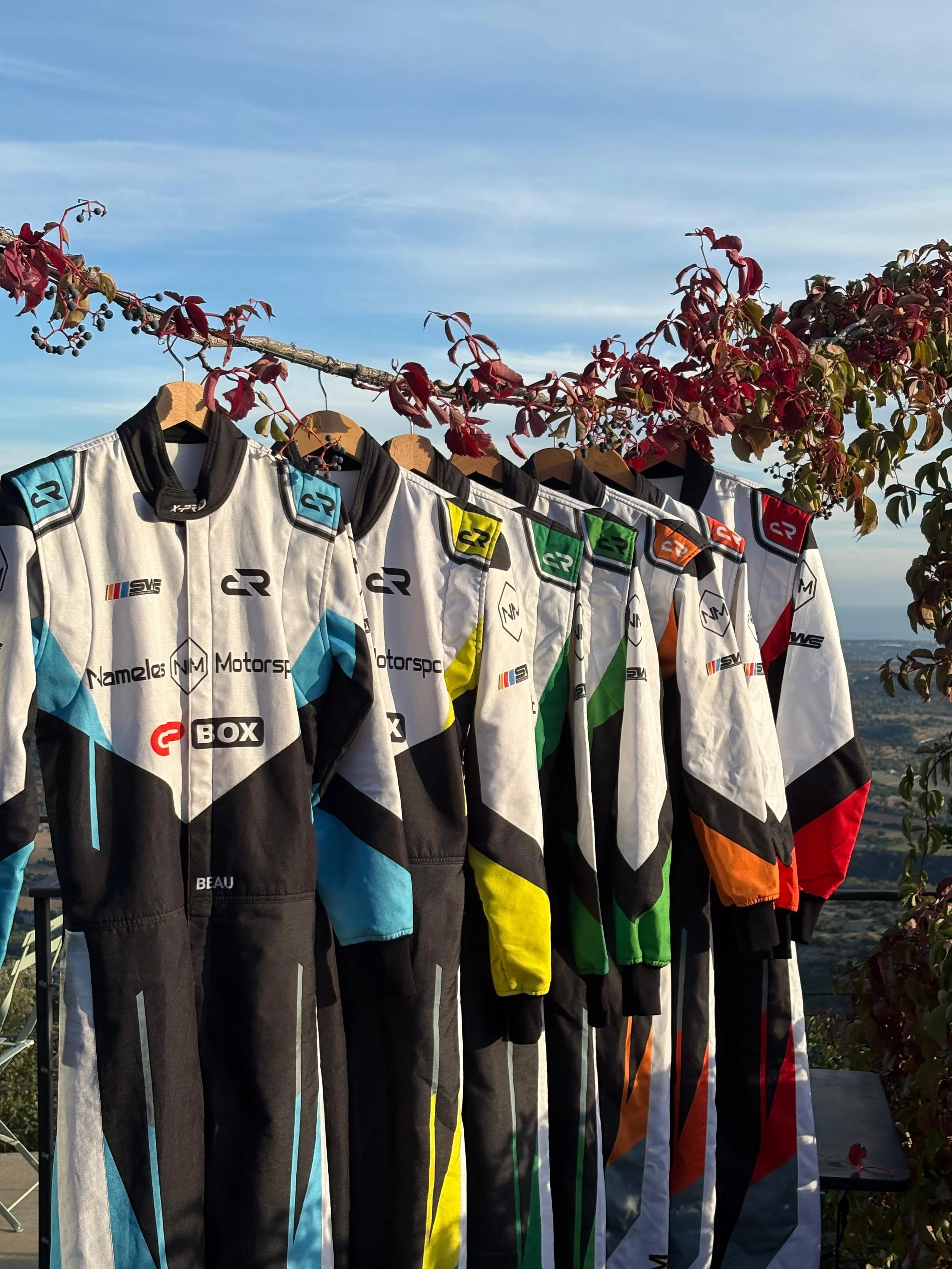 A row of racing suits hanging on a clothesline outdoors with trees and a mountain landscape in the background.