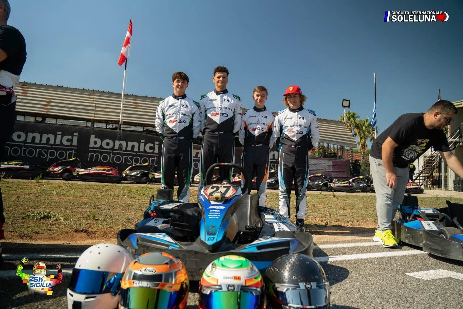 Race car drivers in racing suits stand together at Motorsport race track with go-karts and helmets in the foreground and flags in the background.