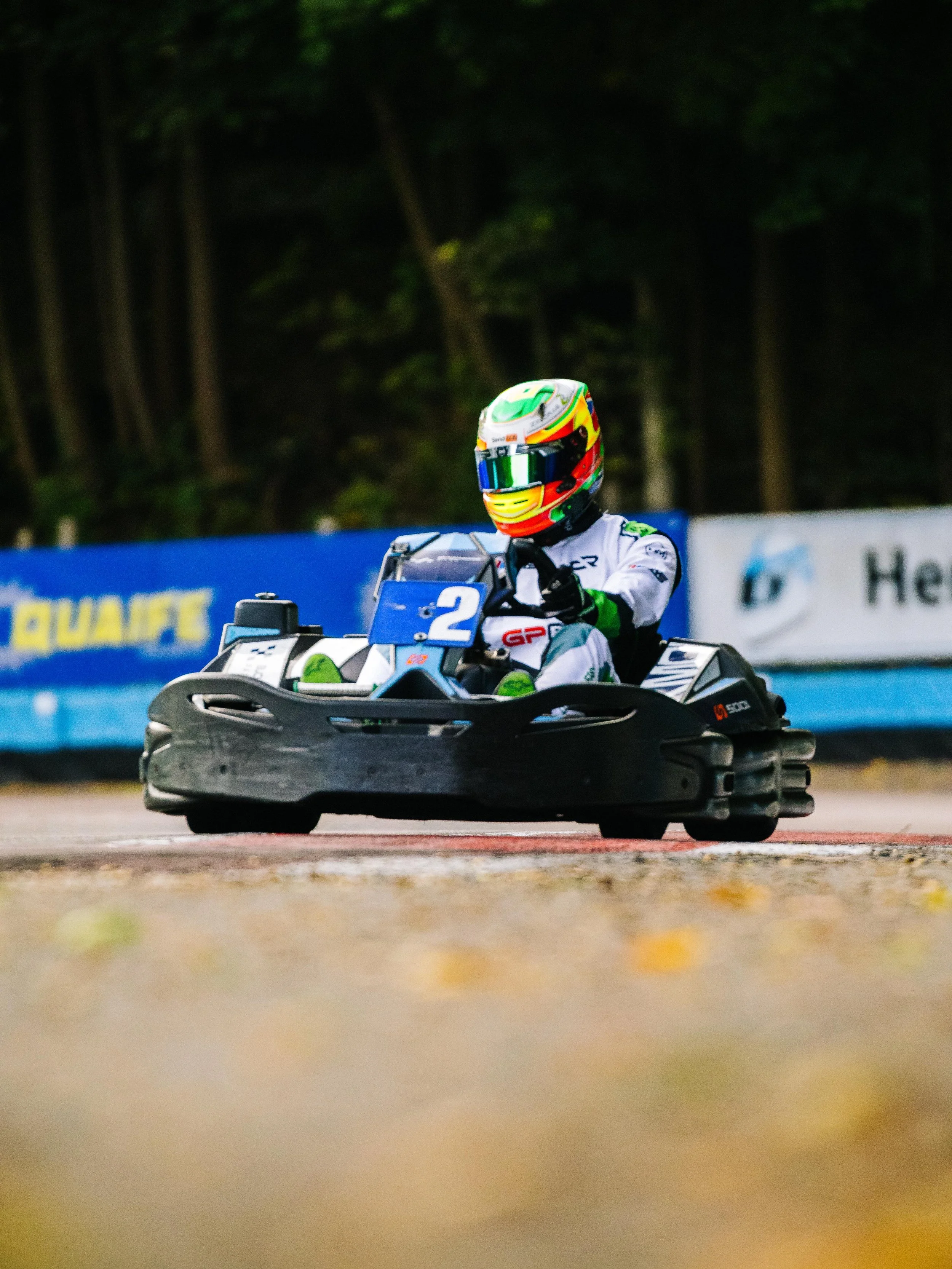A person racing a go-kart on a track, wearing a colorful helmet and a white racing suit, with a forest background and advertisement banners.