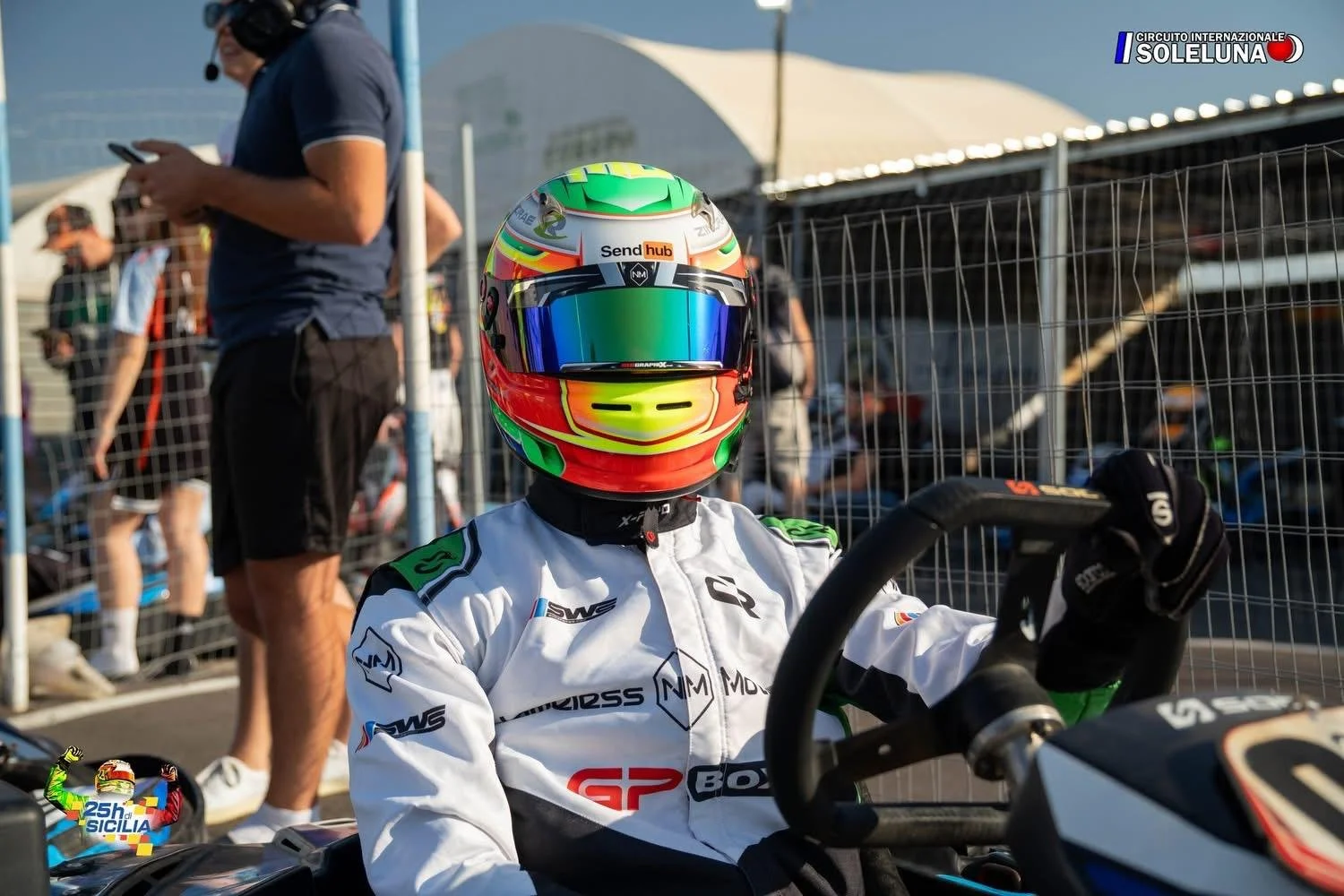 Race car driver in white racing suit and colorful helmet sitting on a race vehicle at the Circuito Internazionale Solleuna, with spectators and security fencing in the background.