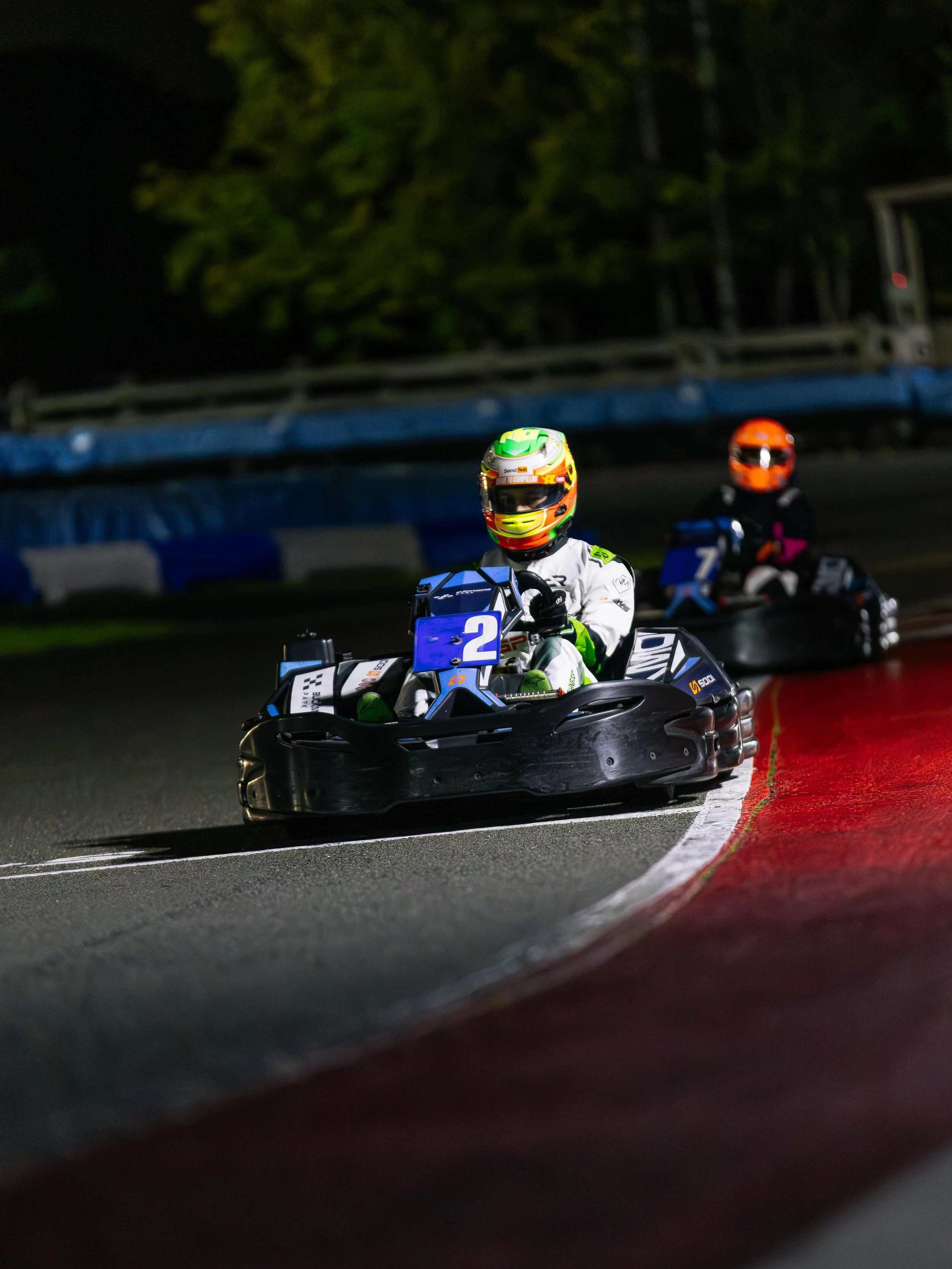 Nighttime go-kart race with two racers wearing colorful helmets and racing suits on a curved track, with trees and barriers in the background.