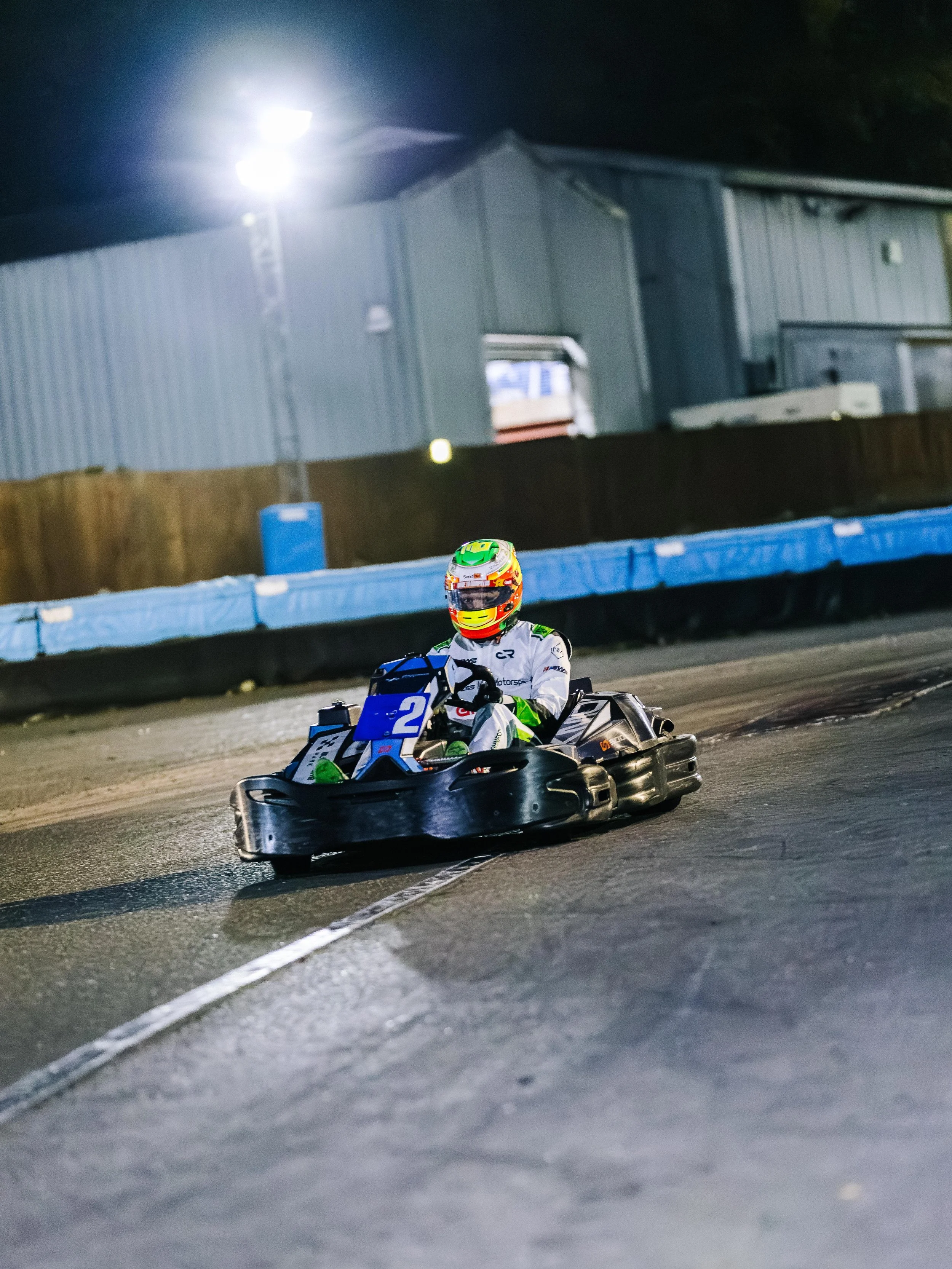 A person riding a go-kart on an indoor track at night, wearing a white racing suit and a colorful helmet, illuminated by bright overhead lights.