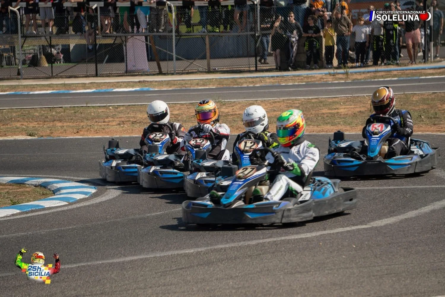 Five go-kart racers wearing helmets and racing suits competing on a track. Spectators watch behind a fence. The track has blue and white curbs, and there is a scenic outdoor setting with grass and trees.