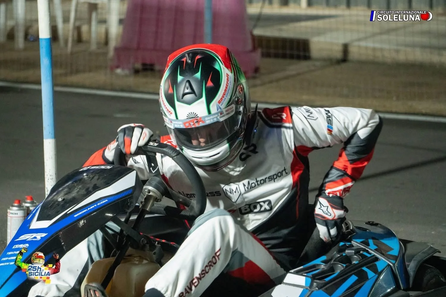 Motorcycle racer adjusting helmet next to a motorcycle at a nighttime racing event, with logos and sponsor decals visible.