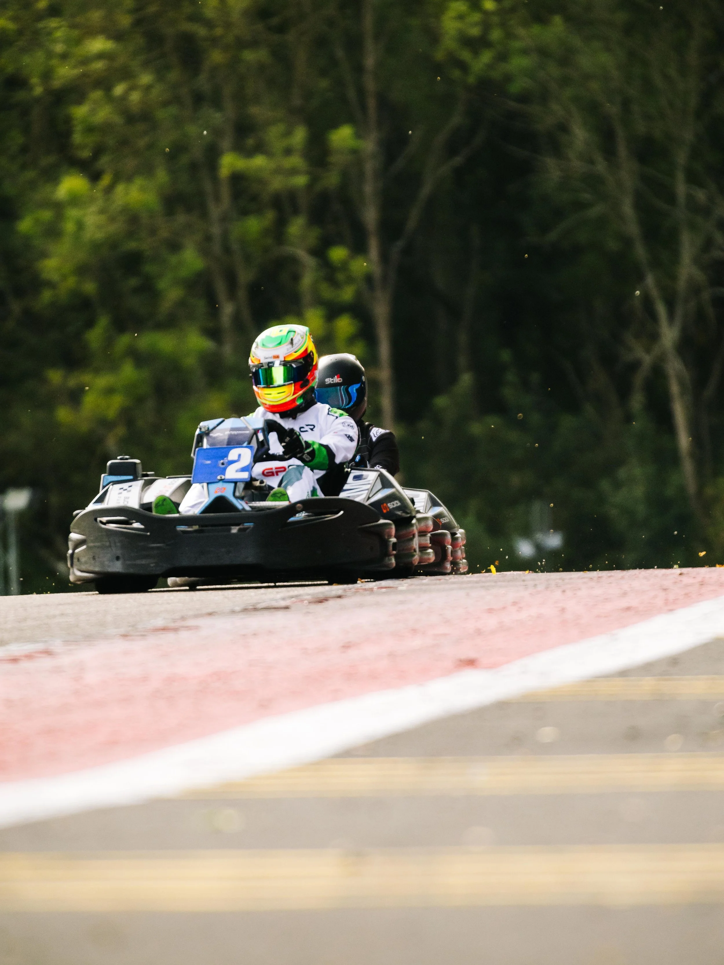 Two people racing in a go-kart on a track with trees in the background.
