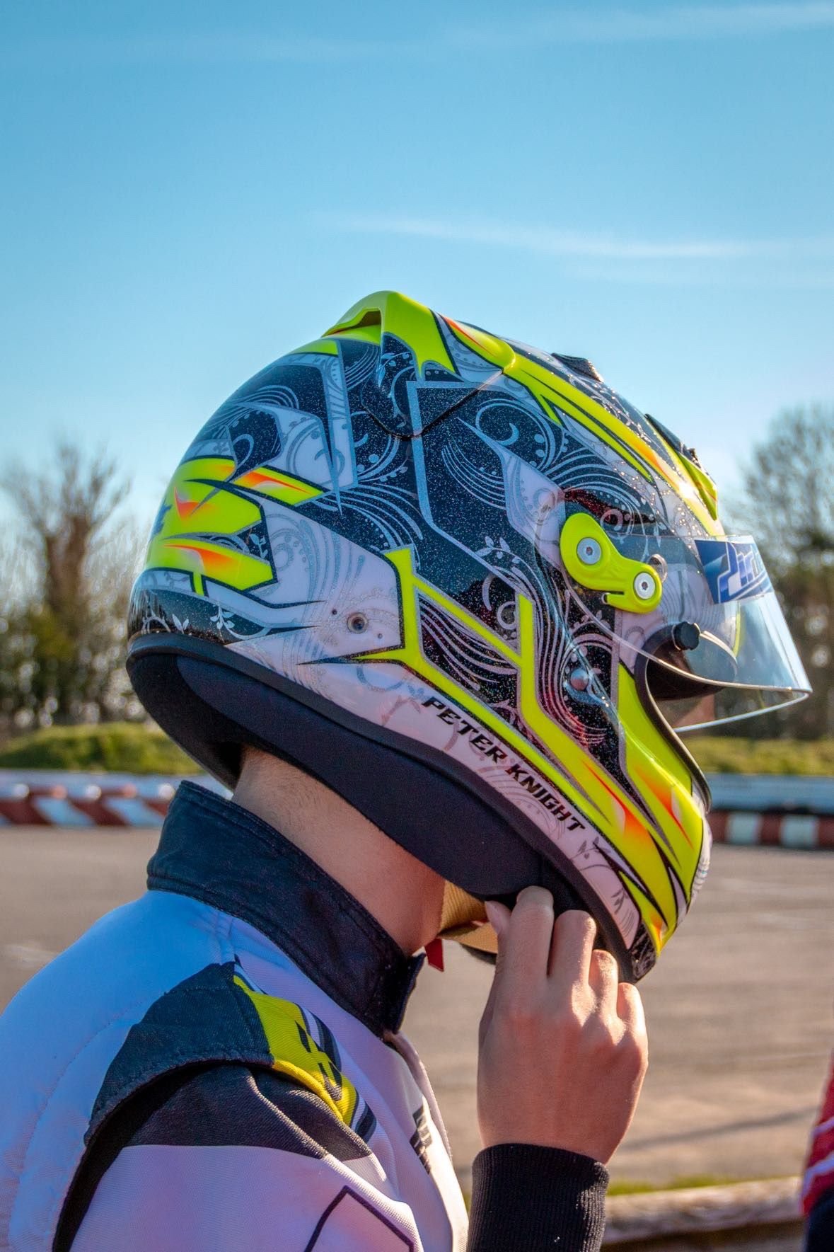 Race car driver wearing a white helmet and racing suit with a prominent logo, standing in a pit area.