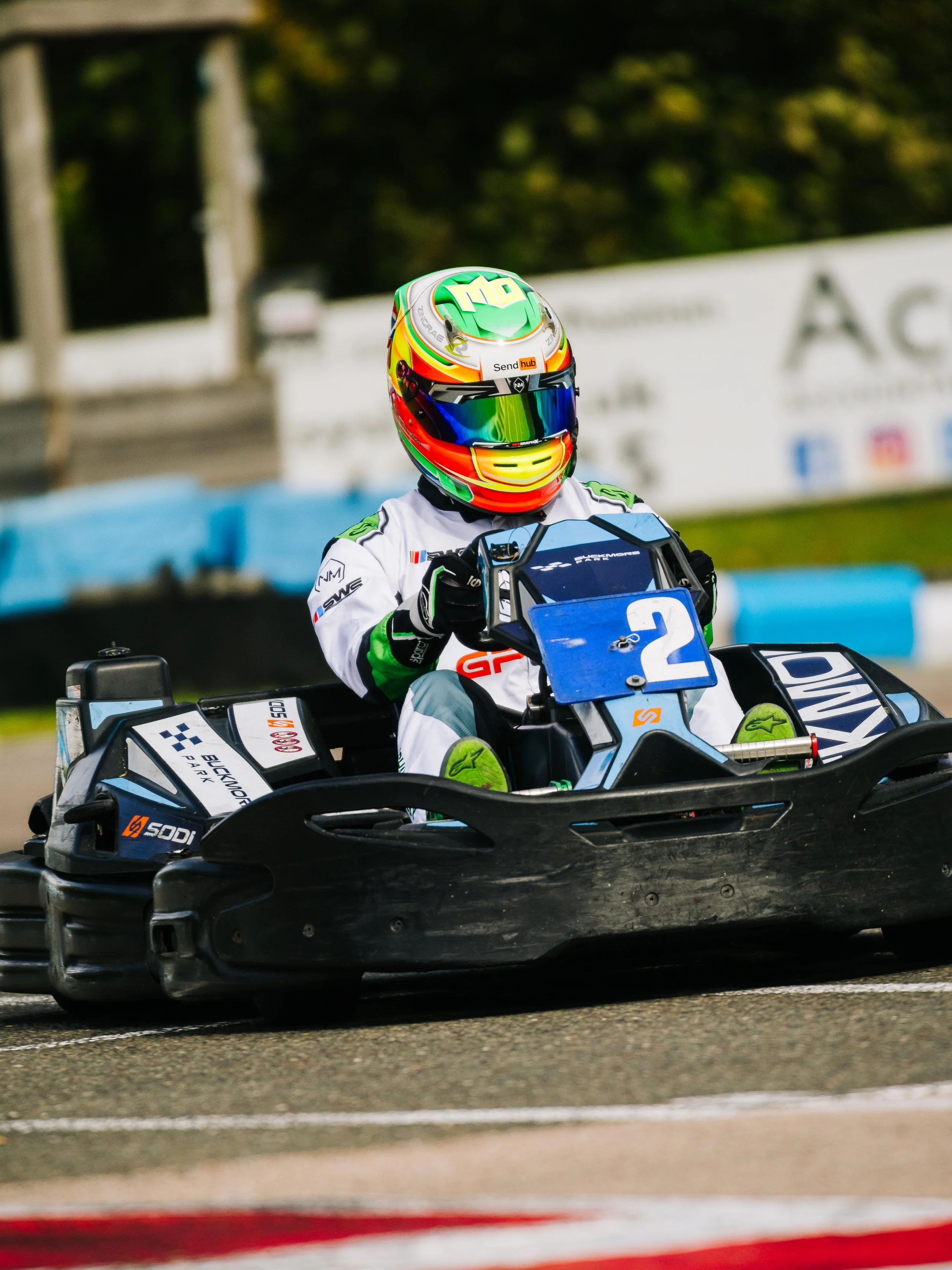 Racecar driver in colorful helmet and racing suit driving a go-kart on a race track.