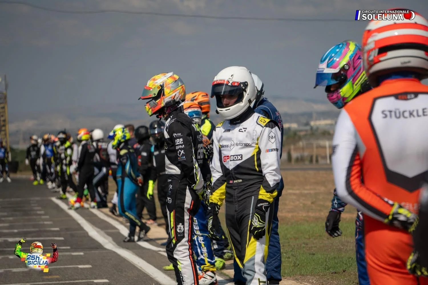 A group of racing drivers standing in a line on a race track, wearing helmets and racing suits, with a cloudy sky and distant landscape in the background. The event is the 25th edition of the Sicily racing race at the Sol LUNA circuit.