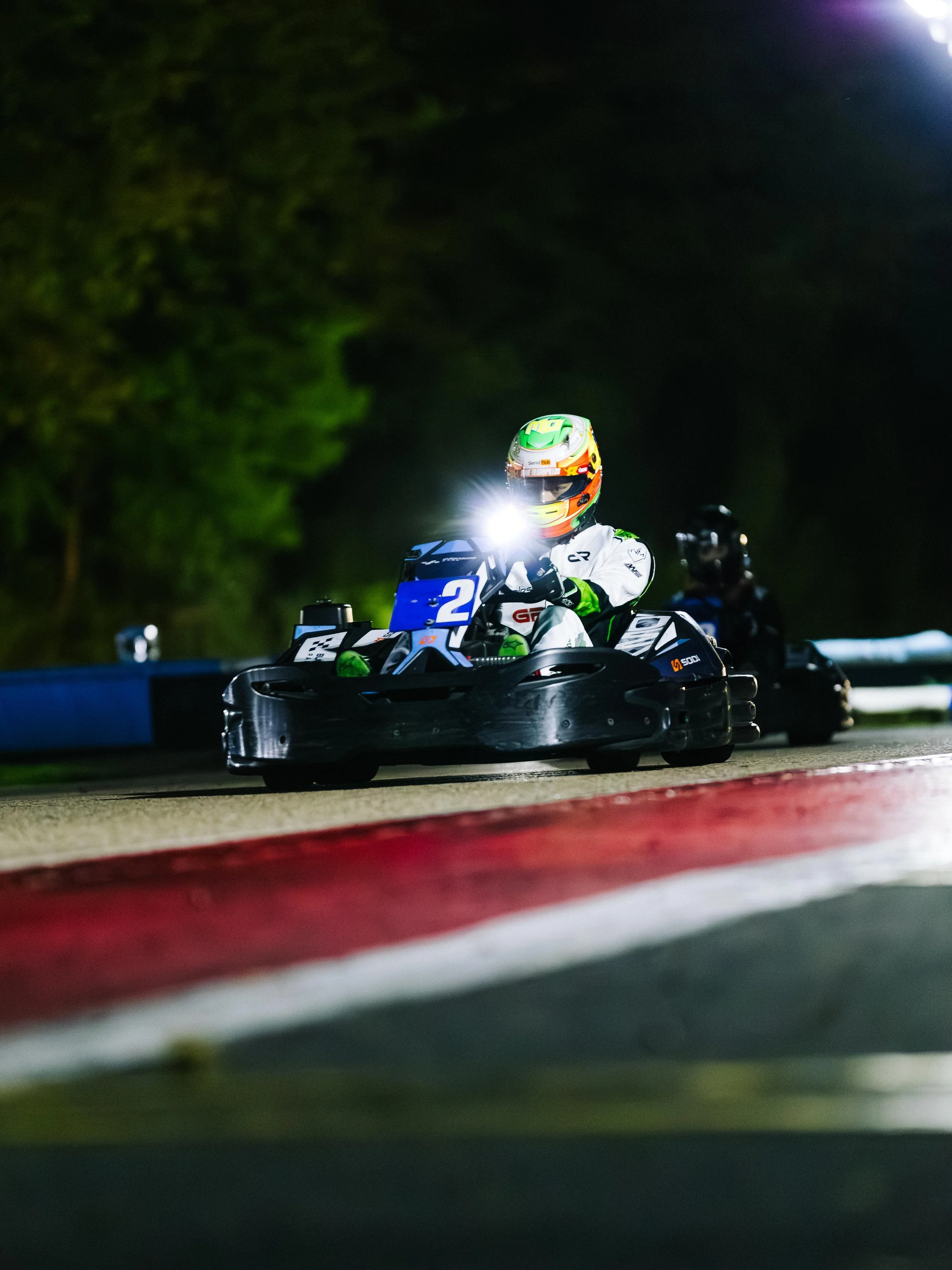 A go-kart racer wearing a helmet and white racing suit on a black go-kart at night, with the kart's headlight on, seen on a race track with red and white curbing.