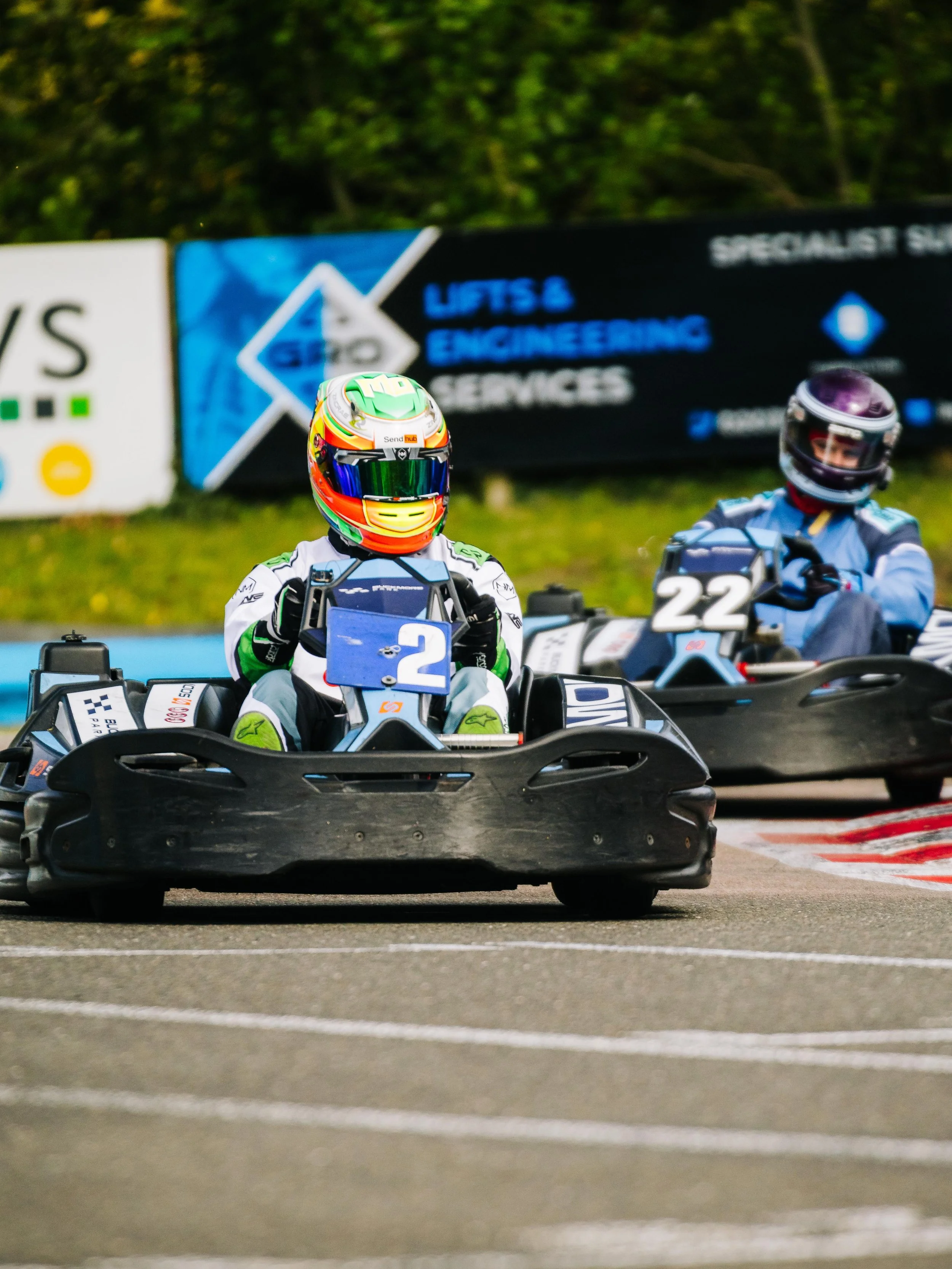 Two go-kart racers on the track, wearing helmets and racing suits, with the number 2 visible on the front of the nearest cart and the number 22 on the second cart in the background.