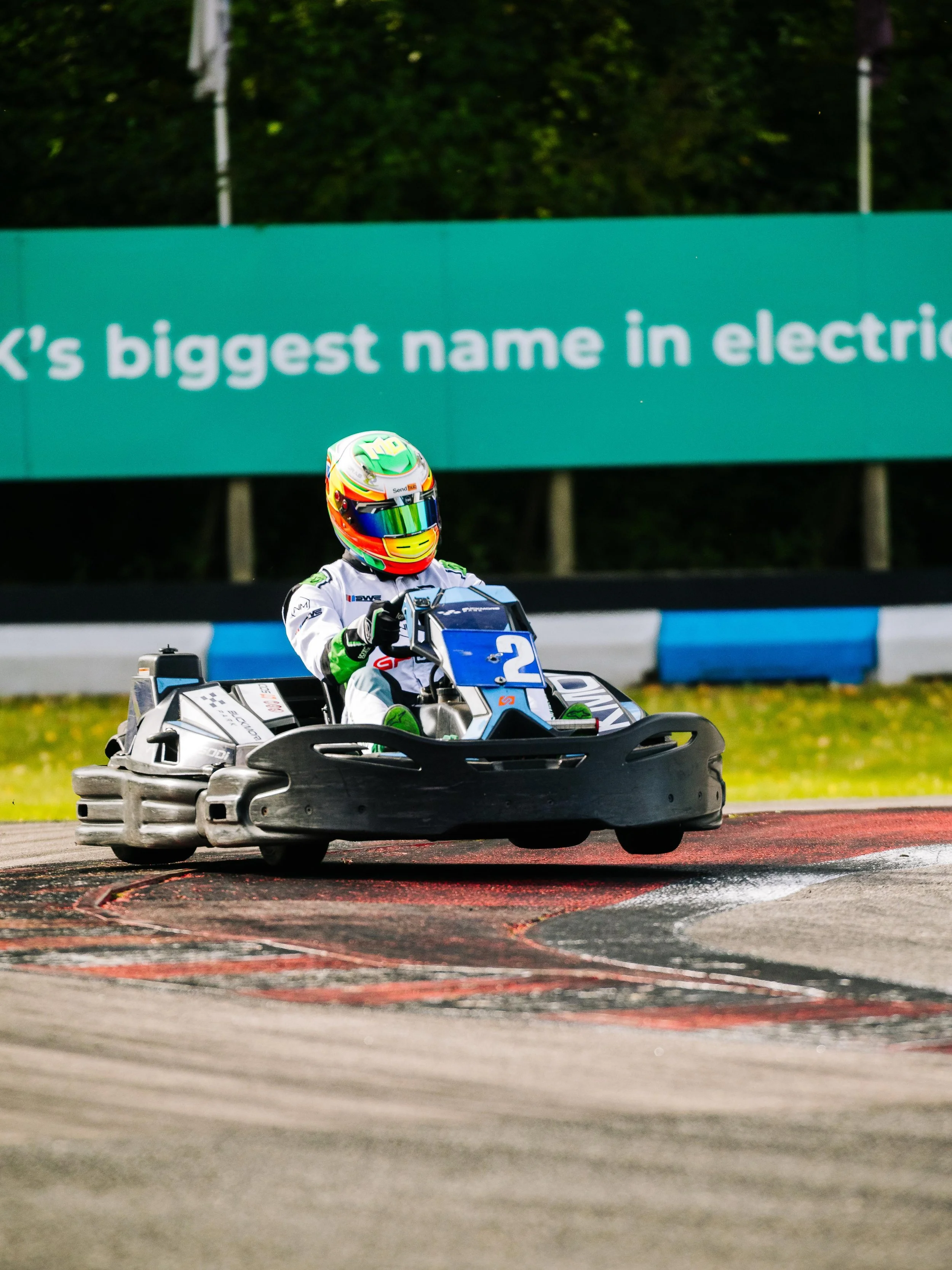 A go-kart racer wearing a colorful helmet and white racing suit driving on a race track.