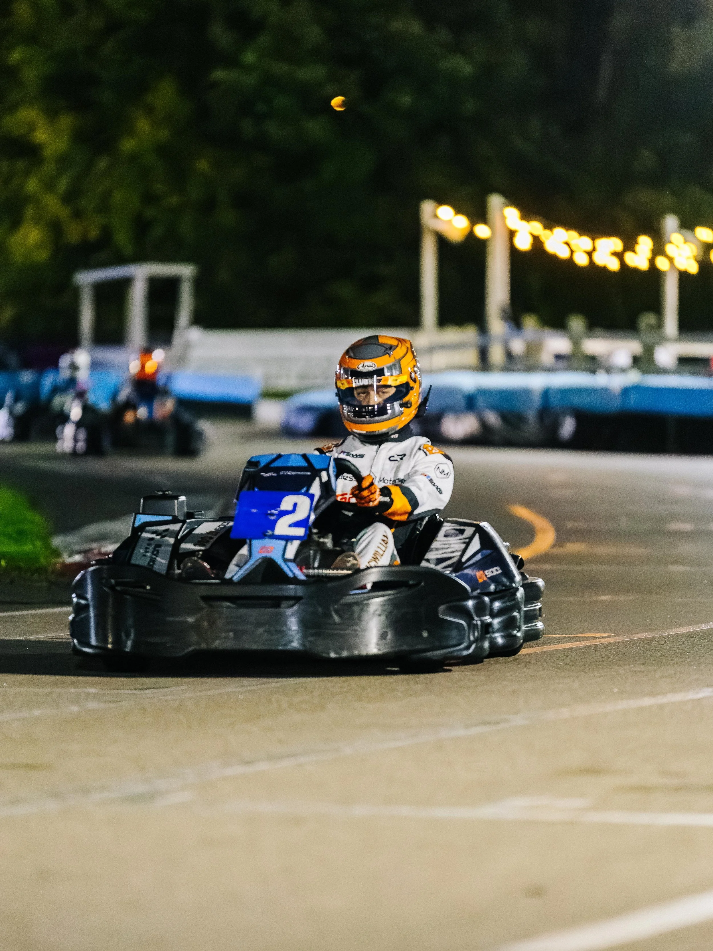 A driver wearing a white racing suit and orange helmet driving a go-kart on a nighttime outdoor track.